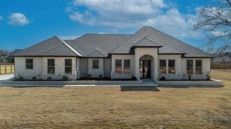 Welcome to 12321 Madison Xing in Bullard, Texas — a truly impeccable new build. This home offers breathtaking curb appeal, showcasing a striking blend of stone and stucco that delivers a modern Texas Mediterranean aesthetic. A beautiful, extended flower garden lines the front of the property, perfectly complementing the architecture and creating a warm, inviting first impression. Step inside through a one-of-a-kind custom foyer that immediately sets the tone for the rest of the home. The oversized living room features vaulted ceilings with exposed beams, anchored by an eye-catching floor-to-ceiling fireplace that serves as the heart of the space. Maple cabinetry throughout the home ties together a thoughtfully curated color palette, creating a cohesive and upscale feel. The kitchen is nothing short of stunning, highlighted by an oversized island with quartz countertops, a farm-style sink, and an impressive boxed ceiling detail that showcases true craftsmanship. A custom vent hood crowns the top-of-the-line 8-burner gas range, perfectly balancing function and design for anyone who loves to cook and entertain. The primary suite is a true retreat, featuring elegant millwork, generous space, and a spa-like bathroom complete with a walk-in shower with multiple shower heads, a standalone soaking tub, glass enclosure, separate vanities for him and her, and an additional makeup vanity. A large walk-in closet completes this private oasis. This home also includes a mother-in-law suite, ideal for extended family or overnight guests, along with two additional bedrooms. A dedicated professional office with custom built-ins and ample storage makes working from home effortless—just bring your computer and get to work. Out back, the flat and spacious backyard is perfect for outdoor enjoyment. Under the covered patio, you'll find a spacious sized breathing room, deal for entertaining all summer long. Call for a tour today.
