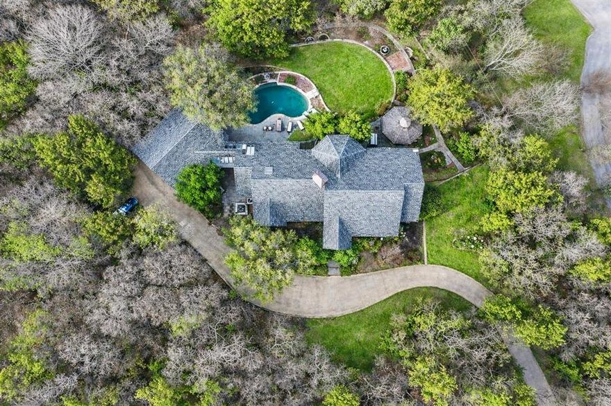 Discreetly nestled on 2.48 private wooded acres in Cedar Hill, this custom 4,000+ sq ft residence offers refined luxury, architectural character, and estate-style privacy. Accessed by a winding drive and framed by stone entry, the home is surrounded by mature trees and serene natural views.
Inside, soaring vaulted ceilings, skylights, rich woodwork, designer built-ins, and wood-framed windows create warmth and architectural distinction. The main level is designed for both entertaining and everyday living, featuring two living areas anchored by stone fireplaces, a formal dining room, and a kitchen with breakfast area overlooking the landscaped grounds and resort-style pool, flowing seamlessly into a wet bar. The primary suite occupies its own private wing and includes a spa-inspired bath with garden tub, separate shower, double vanity, dual walk-in closets, and a distinct adjoining sitting area ideal for lounge, nursery, office, or dressing space. A dedicated home office with adjoining TV room further enhances the flexibility of the main floor. Upstairs, two additional bedrooms and full bath are complemented by a substantial secondary living area with wet bar, creating a private retreat well suited for guests, teens, or multi-generational living. Additional features include a large pantry, spacious laundry room with storage, newer roof, and three AC units. Outdoor living includes a pool, gazebo, expansive patio, and a layered landscape design that frames a defined lawn and gathering areas before transitioning naturally to wooded privacy beyond. Completing the property is a detached three-car garage, separate workshop, and lawn equipment garage. While offering the privacy of an acreage estate, the home remains conveniently positioned near Joe Pool Lake recreation, regional trail systems, major thoroughfares, and access to DFW Airport and the greater Dallas area.