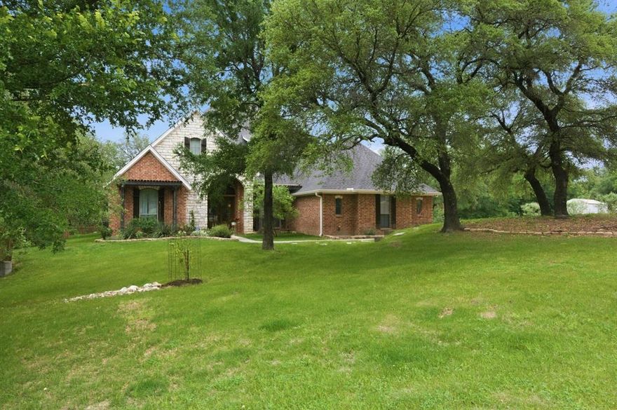 A gated drive leads to this inviting brick home, where a covered entry opens to a bright interior defined by warm wood flooring and thoughtful architectural details. The foyer is anchored by a statement chandelier and a pair of wood-framed glass doors, with an arched opening that draws you into the main living spaces. The main living area offers a comfortable, open layout with a stately fireplace and multiple sets of glass doors that bring in natural light and connect to the outdoors. A secondary living or sitting area provides additional flexibility, while the dining space is set just off the main room for easy flow.  The kitchen features abundant storage in light cabinetry and bar seating with tasteful pendant lighting overhead. Stainless steel appliances, including a double oven, complement the clean design, while a tiled backsplash and ample counter space enhance functionality. The primary bedroom showcases wood flooring, a distinctive wood-beam ceiling, and room for a sitting area. The en suite bath includes a freestanding tub, a separate tiled shower, and a dual-sink vanity, along with a generously sized closet outfitted with built-in organization. Outdoors, a screened porch offers a relaxed setting with views of the surrounding greenery. The backyard includes a raised, terrace area overlooking the rest of the grounds, with wooden railings, a paver walkway, a fire-pit, and broad stairs leading down.  Enjoy plentiful mature trees, a convenient metal outbuilding, and lots of room to relax and entertain, all fully fenced.  Easy access to Hwy 199 and nearby spectacular Eagle Mountain Lake with fishing, boating, trails, parks, and more.