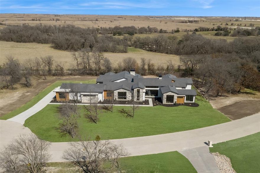 Tucked into a cul-de-sac in the gated Rancho Ladera, this home is a masterfully crafted estate by renowned builder Sean Knight, with 6,333 sq. ft of refined living on 2.147 acres. The steel, stone, and white oak contemporary home features 4 bedrooms, 4 full baths, 2 half baths, all designed with elevated finishes.
A dramatic glass and steel front door opens to a striking entry with a dramatic chandelier and white oak ceiling. The expansive living room has a floor-to-ceiling quartzite hearth and gas fireplace, creating a focal point for gatherings large and small. Refrigerated wine storage is a collector’s dream. The chef’s kitchen showcases Thermador appliances, quartz countertops, a breakfast nook, and a dedicated catering kitchen. A mudroom opens to the driveway, while an oversized laundry room provides extensive cabinetry and workspace.
The split-bedroom layout ensures privacy. The primary suite is a spa-inspired retreat with private courtyard access, gas fireplace, steam shower, water room, marble tile, concrete soaking tub, beverage refrigerator, a custom double closet complete with stacked washer, dryer, and steamer. Secondary ensuite bedrooms include a guest suite with private bath and a bunk room sleeping 4-6, with toy storage concealed behind a custom bookcase. A dedicated office with fluted paneling hides a walk-in closet.
Designed for recreation, the home features a media-game room with patio access and a wet bar highlighted by a dramatic backlit onyx bar top. An exercise room with half bath and a secure tornado shelter add convenience and peace of mind. The 4.5-car garage includes epoxy flooring and a custom car lift.
Outdoor living rivals the interior with a covered patio, fireplace, and Coyote grill kitchen. With room for a future pool and frequent visits from deer and wild turkey, the setting feels both private and serene. Located in highly acclaimed Aledo ISD and minutes from Bluejack Ranch, residents enjoy luxury living and in a prime location.