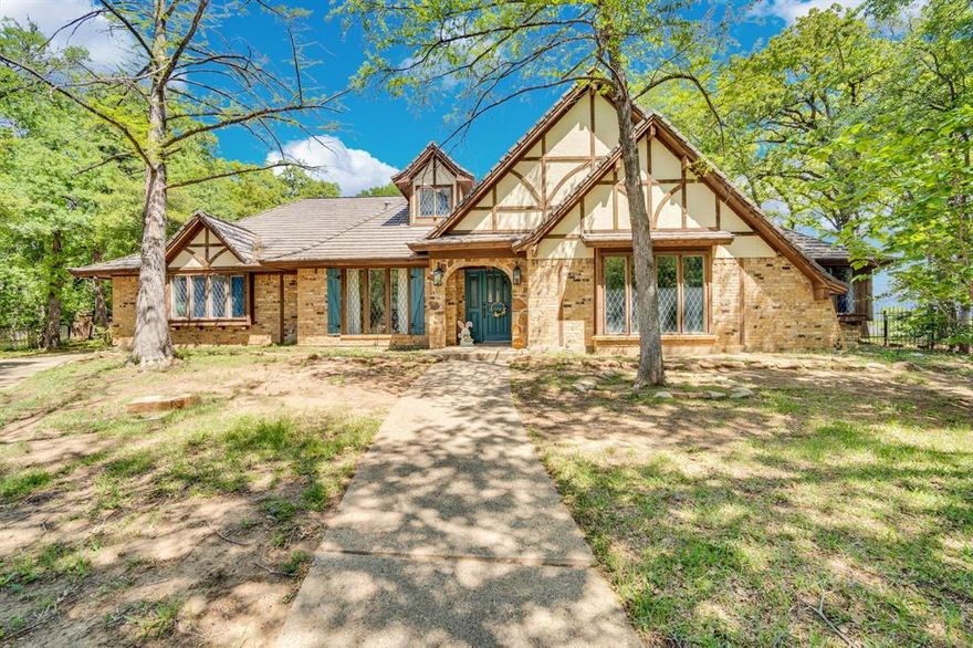 Nestled beneath a canopy of mature trees, this stunning Tudor-style home feels like a storybook French cottage tucked away in the heart of Fort Worth—yet just minutes from Arlington’s Entertainment District and Downtown Fort Worth. Built in 1976 and rich in character, this expansive 3,863 sq ft home offers 4 bedrooms, 3 full baths, 1 half bath, 3 living areas, and 3 fireplaces—perfect for a large family or anyone who appreciates timeless charm.
The main floor showcases beautiful hardwood flooring, a formal dining room, and a spacious galley-style kitchen with granite countertops, hardwood cabinetry, stainless steel appliances, and a wide peninsula ideal for prep and gathering. Two primary suites are conveniently located on the first floor, making this home ideal for multigenerational living. One of the suites has been fully updated with a luxurious soaking tub and spa-like finishes. A private office on the first floor can easily function as a fourth bedroom, and the oversized, newly updated laundry room offers abundant storage.
Upstairs features a large game room or man cave complete with a wet bar and a beautifully updated half bath. An elevator provides convenient access from the first floor to the upper level—adding both comfort and functionality. The oversized family room is warm and inviting, centered around a grand fireplace and filled with natural charm. Step outside to your private backyard retreat featuring a full outdoor kitchen with sink, gas grill, burner, sitting area, outdoor chimney, and hot tub—perfect for entertaining. A separate powered bonus space, offers endless possibilities as a home office, gym, or creative space. The fully fenced yard and oversized 3-car garage complete this one-of-a-kind property. A rare opportunity to own a home full of character, space, and unique features in an unbeatable Fort Worth location with quick access to everything.