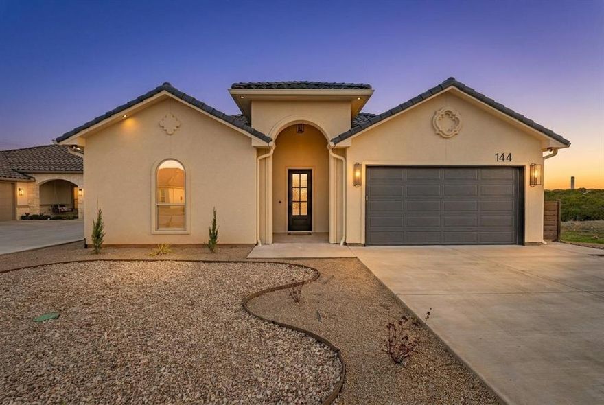 Experience luxury living with breathtaking Hill Country views in this exceptional new construction home by BellaVue Homes, located in the desirable Tuscan Village community of Glen Rose, Texas. Designed with elegance and comfort in mind, this 3-bedroom, 2-bath residence showcases soaring 14-foot vaulted ceilings, abundant natural light, and a thoughtfully designed open-concept floor plan. The spacious living, dining, and kitchen areas are anchored by a striking stone fireplace with electric insert and expansive windows that frame the scenic surroundings. The kitchen features granite countertops, abundant cabinetry, generous prep space, and a large center island—perfect for entertaining and everyday living. Unwind in the private primary suite, complete with oversized windows and tranquil views. The spa-inspired ensuite bath offers dual vanities, a garden tub, a walk-in shower, and a massive walk-in closet with convenient access to the utility room. The oversized utility room includes a sink, built-in cabinetry, and a dedicated workspace ideal for a home office or craft area. Two generously sized guest bedrooms share a well-appointed full bath. Enjoy peaceful evenings and spectacular Texas sunsets from the expansive covered back patio. Exterior highlights include Spanish-style stucco, durable tile roofing, and low-maintenance desert landscaping. Residents of Tuscan Village enjoy access to premium amenities including a community clubhouse, swimming pool, fishing pond, and scenic walking trails. This home offers the perfect blend of Hill Country charm, modern design, and resort-style community living.