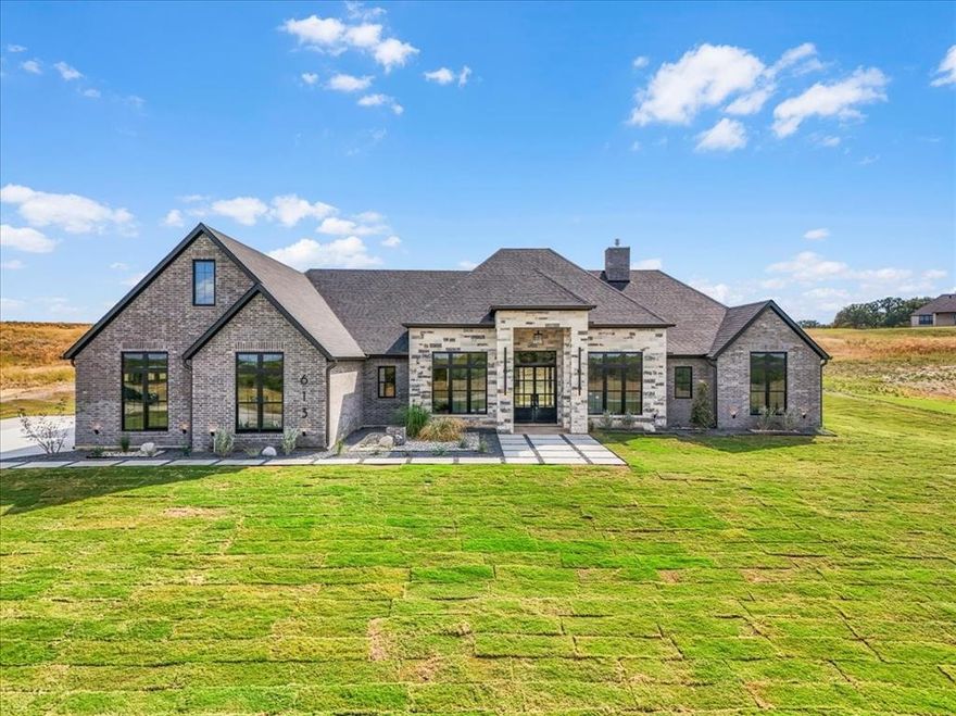 Builder offering interest rates as low as 5% for qualified buyers—an incredible chance to lock in luxury at a lower rate! Set on a 2.35-ac lot within Brock ISD’s premier gated community, this residence blends sophistication and serenity. Through custom steel double doors framed by modern vertical pillar lights, guests enter a showstopping foyer. The formal dining room stuns with wainscoting, while the executive office features built-ins and French doors for refined functionality. At the heart of the home, a chef-inspired kitchen designed for entertaining impresses with a Taj Mahal quartzite waterfall island, counters & backsplash, white oak inset cabinetry with black hardware, and commercial-grade appliances. A secondary prep kitchen with oven, microwave, sink & patio access ensures effortless hosting. The great room, anchored by a floating fireplace, features contemporary fixtures and pillar lighting that infuse a warm, modern glow throughout. Engineered white oak flooring flows through the main living areas beneath 16-ft ceilings, adding natural warmth to the home’s sleek design. Expansive glass sliders open to a cathedral-style covered patio with stained tongue-and-groove ceiling—ideal for evening gatherings under Texas skies. The primary suite delivers spa-like tranquility with a circular soaking tub, glass-enclosed rainfall shower, dual vanities, and two oversized walk-in closets. A private sit-down vanity connects gracefully within the suite, blending beauty and function. Three additional bedrooms each offer ensuite baths and walk-in closets. Behind the kitchen, a staircase leads to a private loft with full bath, closet & attic access—perfect for a gym, media room, or creative retreat. Outdoors, enjoy xeriscape landscaping with natural boulders, a buried propane tank, private well, aerobic septic, and the rare freedom to keep horses or chickens. M2 Luxury Homes once again delivers a residence that defines elevated living and modern Texas luxury.