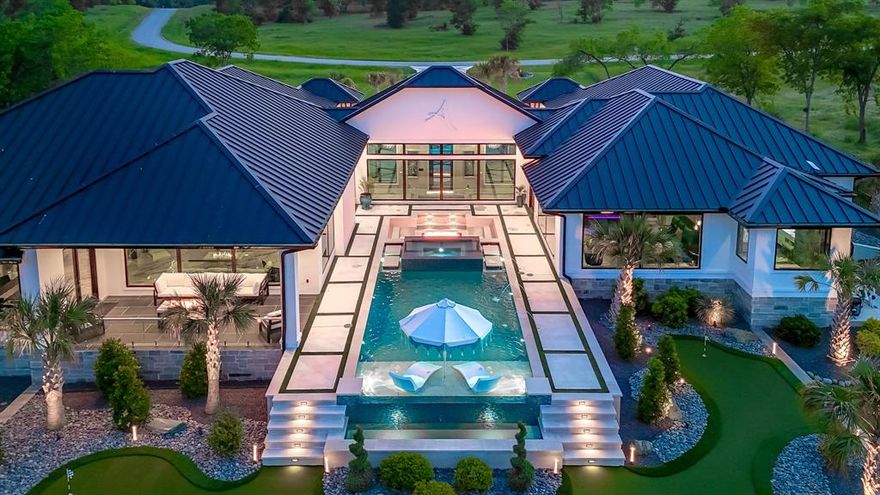 Modern luxury meets lakeside living in this stunning 4-bedroom, 4-bathroom home on just over an acre of waterfront property. Designed with clean lines and top-tier finishes, this home offers a perfect balance of elegance, comfort, and functionality. Arrive in style on a turf-inlay driveway leading to a 4-car garage with epoxy flooring, heated and cooled climate control, and a modern glass door. Inside, the open-concept living space features large windows with breathtaking views of the lake and heated infinity pool, plus an electric fireplace for cozy ambiance. The chef's kitchen is equipped with a massive island, built-in granite sink, brass refrigerator and freezer, Scotsman ice maker, and a walk-in pantry. Entertain with ease thanks to the wet bar located in one of the bedrooms. Retreat to the luxurious primary suite with an electric fireplace, spa-inspired ensuite bathroom with double vanities, garden tub, separate shower, and a custom walk-in closet. The dedicated office features sleek glass walls, perfect for working from home in style. Step outside to tile-covered porches and patios, glass railing for unobstructed views, and a porcelain tile fire pit for relaxing evenings. Enjoy two putting greens, and a custom boat lift with remote control- designed for effortless lake access. With a heated LED-lit infinity pool and a seamless indoor-outdoor flow, this property is a rare blend of sophistication, comfort, and lakeside serenity. Don't miss your chance to own this fully customized modern retreat.