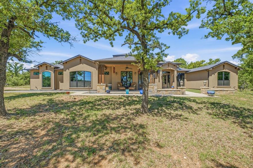 Nestled on 43 breathtaking acres, this stunning stucco ranch-style home offers 2,384 sq of thoughtfully designed living space, blending rustic charm with refined luxury. A long, private driveway framed by wooden fencing sets the tone for the peaceful country retreat that awaits. Step inside to a grand living area where soaring ceilings, exposed wood beams, & a striking floor-to-ceiling stone fireplace create a warm yet sophisticated gathering space. Large windows invite natural light & showcase the surrounding landscape, bringing the outdoors in. The kitchen is truly a chef’s dream, featuring a commercial grade gas cooktop, double oven, a wine fridge & an expansive center island perfect for entertaining. Custom cabinetry, abundant counter space & a spacious walk-in pantry with additional wine storage make this kitchen as functional as it is beautiful. The private master suite offers a luxurious escape, complete with a spa-like en-suite bath featuring dual vanities, a dedicated makeup area, an oversized tiled shower & a generous walk-in closet. A split-bedroom layout provides added privacy, with well-appointed guest bedrooms that each include walk-in closets & share a stylish hall bath. Outdoor living is just as impressive, with inviting front & back patio spaces ideal for relaxing or entertaining while overlooking the rolling acreage. The fully fenced backyard, enhanced by a stone retaining wall, offers both security & charm. Additional amenities elevate this property even further, including a workshop with electricity & roll-up doors, a 3000 gallon water storage tank, a generator connection switch for both the home & well house, & two RV hookups—perfect for guests or extended stays. The land itself is a true highlight, featuring lush pastures, mature trees, & two scenic tanks that add to the property’s natural beauty. Whether you’re seeking tranquility, functionality, or a place to entertain, this exceptional property delivers the ultimate country lifestyle.