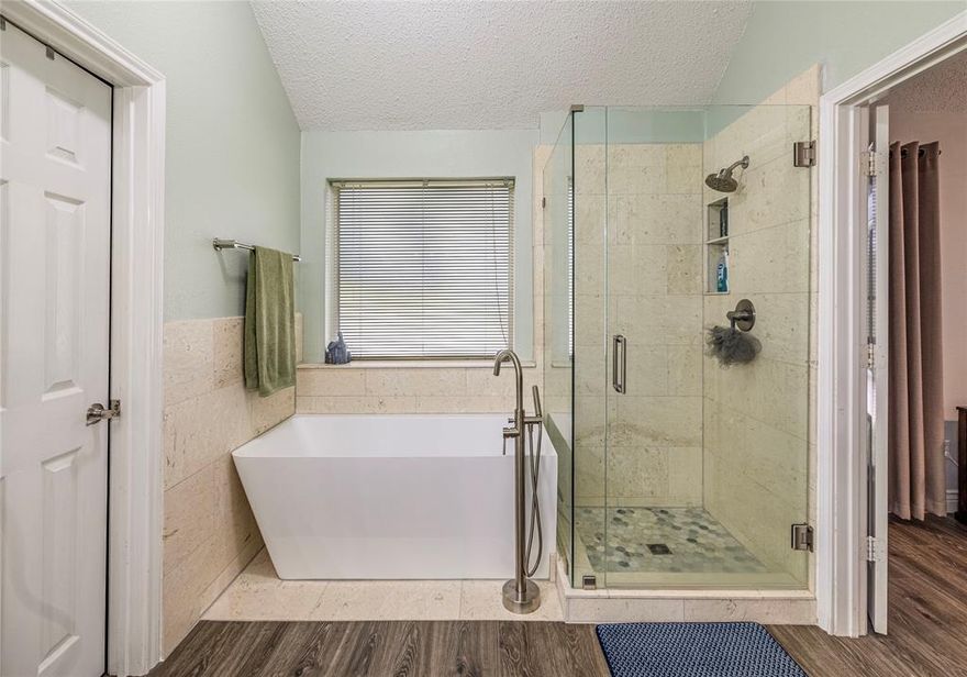 Set on a corner lot with mature landscaping, this home immediately offers a sense of openness—from its tall, welcoming elevation to the easy flow of the interior spaces. Inside, abundant natural light filters through wide windows, guiding you through rooms designed to feel both connected and comfortable. The kitchen blends warmth and function with granite surfaces, a generous island, and richly grained cabinetry, opening seamlessly to the breakfast area and a living room centered around a stone-faced fireplace. Wood-look flooring and gentle architectural arches add texture and continuity throughout the main level. A quiet flex space near the front of the home provides a comfortable setting for focused work or creative pursuits, while the formal dining area is well suited for gatherings and special occasions. The first-floor primary suite offers a peaceful retreat, highlighted by a modern bath with a freestanding soaking tub and a glass-enclosed shower that creates a spa-like atmosphere. Upstairs, an expansive game room invites a variety of uses—from movie nights and music to fitness or play—while nearby rooms adapt easily to changing needs over time. Outside, the covered patio extends daily living with ceiling fans, wide tile flooring, and views of a deep, sun-filled backyard accented by raised planting beds. Whether grilling, relaxing, or enjoying the changing seasons, the space feels both functional and serene. With its flexible layout, inviting outdoor areas, and a location convenient to parks, schools, and major routes, this home offers a setting designed to evolve with the way you live.