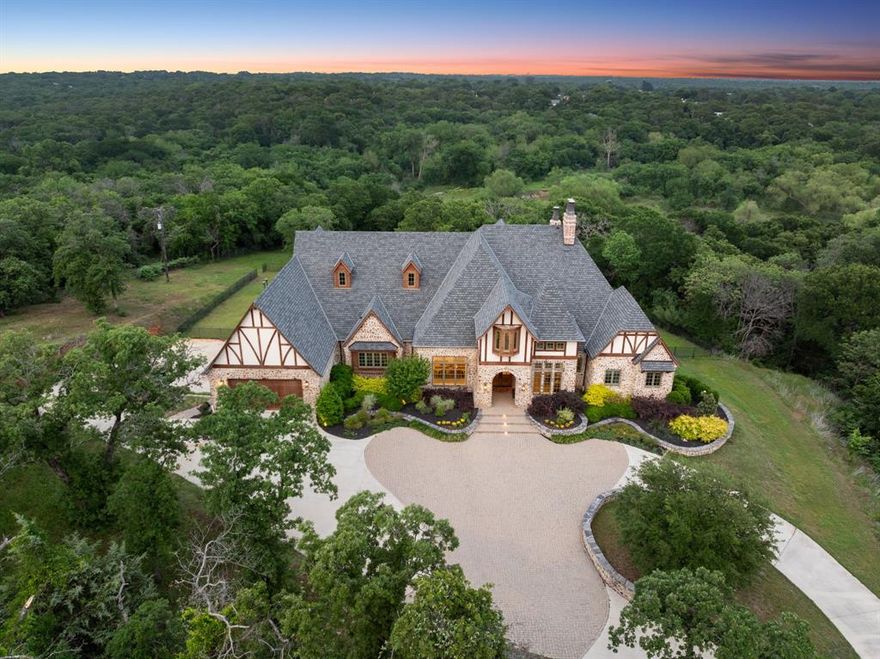 Newly introduced at $4,749,000, this exceptional custom estate sits on 4.5 serene acres, backing to Corps of Engineers land for lasting privacy- offering rare seclusion in the heart of Flower Mound. A gated, tree-lined drive leads to a stunning villa inspired by the English countryside, thoughtfully designed for both everyday living and easy entertaining.
Built into the hillside, this unique home features a full level basement walk-out to the pool and spa, complete with two ensuite bedrooms, enormous entertaining space with game room, gym, wet bar, and dog grooming room. Upstairs, 20-foot beamed ceilings highlight the open-concept living area, anchored by a gourmet kitchen, dining space, and dual offices- all opening to a spacious covered veranda with a stone fireplace and outdoor kitchen, an ideal setting to relax and take in sweeping sunset views.
The primary suite offers a private terrace complete with a fireplace, offering direct access to the lower level outdoor backyard. Additional highlights include a large main-level ensuite guest room, a half court regulation basketball gym which can easily double as a pickleball court, Control4 home automation, oversized laundry and pantry spaces, foam insulation, Cat 5+ wiring, and a Class 4 roof. Room to expand with up to 1,600 additional sq ft. The 3-car garage currently serving as the basketball half court can easily be converted back, making for 5-car garages total. Sweeping circular drive provides ample extra parking.