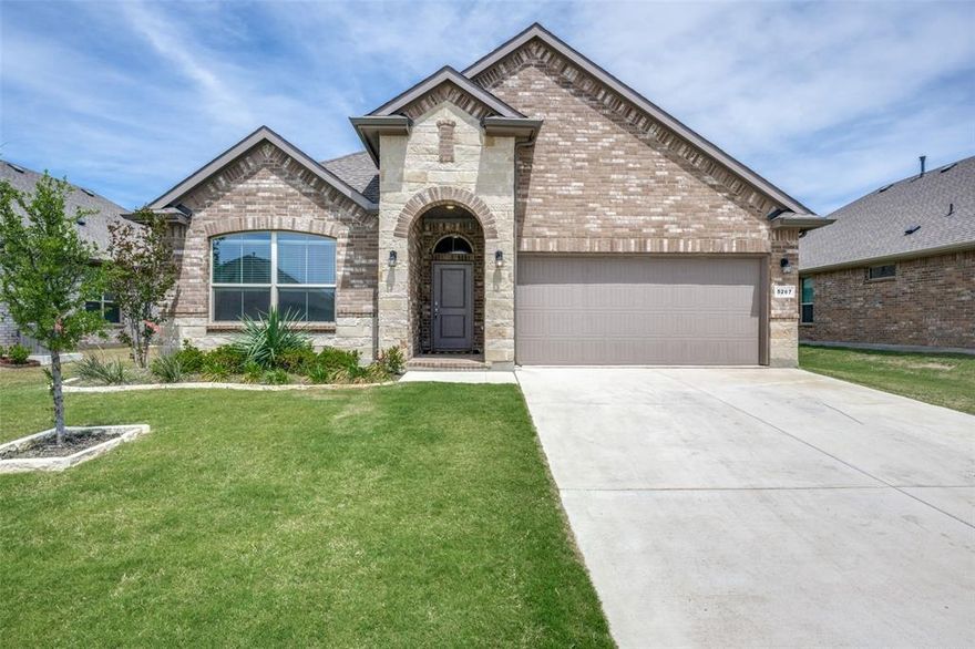 Welcome to this beautifully maintained single-story home offering a bright, open layout and modern finishes throughout. The inviting exterior features a blend of brick and stone, a covered entry, and clean landscaping that sets the tone for the rest of the home. Inside, you’re greeted by warm natural light, neutral colors, and durable wood-look tile flooring that flows through the main living areas.  The open-concept kitchen and living space is the heart of the home, featuring light gray cabinetry, stainless steel appliances, a gas range, a large island with seating, pendant lighting, and a spacious dining area. The living room offers generous windows, a ceiling fan, and plenty of room for comfortable everyday living or entertaining.  The private primary suite includes a large bedroom, walk-in closet, and a stylish ensuite bath with dual sinks, a wide vanity, and a glass-enclosed shower with tile surround and built-in bench. Additional bedrooms are well-sized with neutral tones, soft carpet, and great natural light—perfect for family, guests, or a home office. A built-in desk nook adds convenient workspace, while the separate laundry room and attached two-car garage provide practical storage options. Step outside to a fully fenced backyard with a covered patio, lush lawn, and room to play, garden, or relax. Located in Krum ISD with easy access to nearby amenities, this home blends comfort, function, and modern style in a peaceful neighborhood setting.