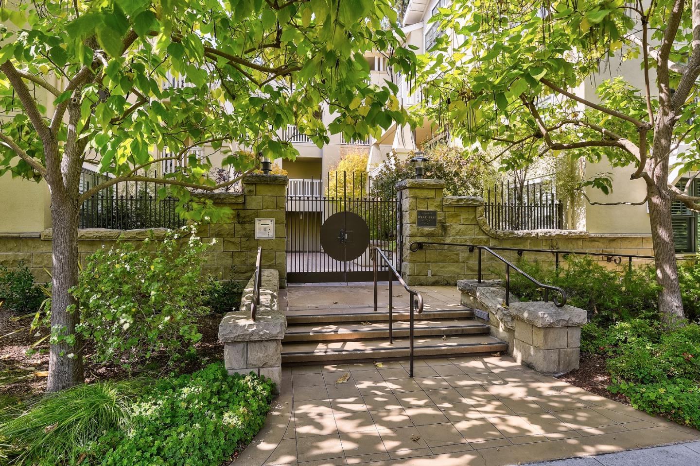 Residential building entrance with a black gate, stone pillars, paved steps, and leafy trees.