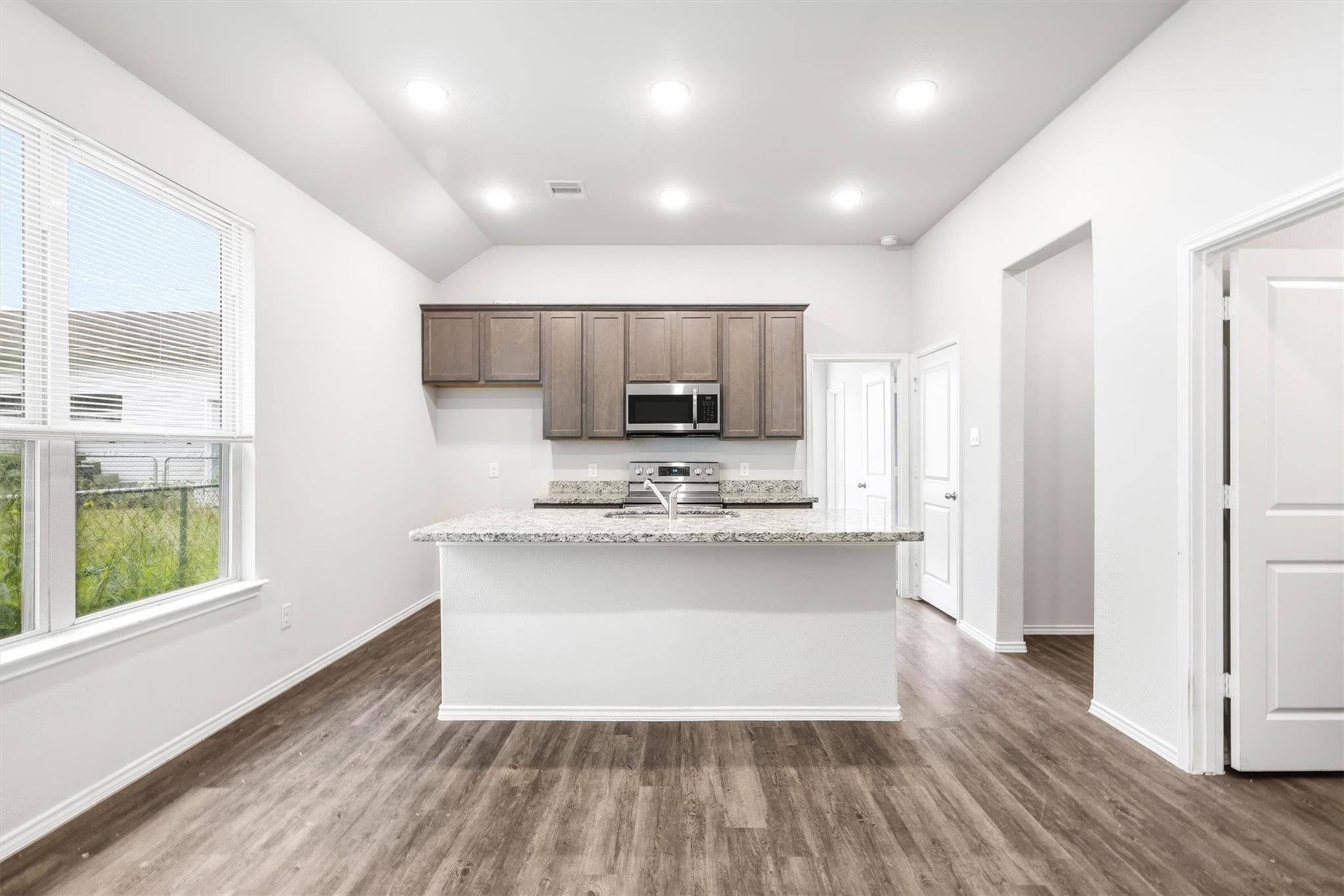 This angle shows just how much natural light pours into this space ? those low-E windows along the left wall keep things bright without the heat, and the vaulted ceiling adds volume that makes 800 square feet feel genuinely comfortable. The granite peninsula with sink sits center stage, with the stainless range and cabinetry lined up cleanly behind it.