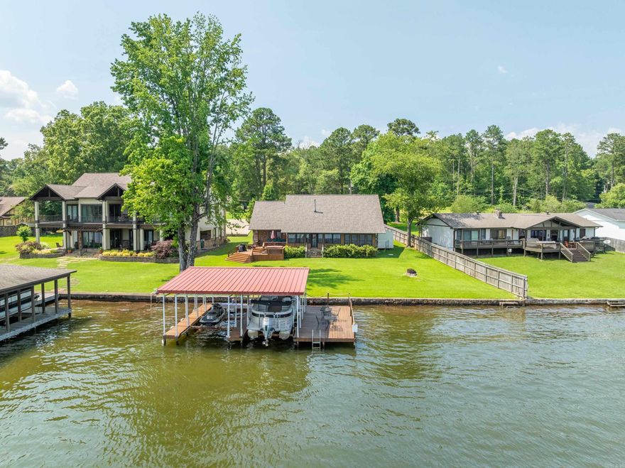 Experience rustic charm and lakefront living in this one-level cabin-style home nestled on the shores of Lake Hamilton. Situated on a truly level lot, this property offers effortless access to the water—just steps from your back door to the double covered dock with a swim deck. Inside, you’ll find 3 bedrooms, each with its own attached bath, plus a versatile flex space currently used as 4th bedroom. You'll love the spacious living area with warm wood beams, and a cozy stone fireplace as the focal point. The highlight of the home is the Arkansas room—an enclosed sunroom spanning the back of the house with expansive windows that capture breathtaking lake views, while allowing you to relax in year-round comfort with heat and A/C. Step outside to a tiered deck perfect for entertaining, complete with a hot tub to relax in while encompassed by water views. Brand-new roof was recently installed (2025), and an in-ground sprinkler system keeps the yard lush. Whether you're looking for a peaceful retreat or a place to gather with friends and family, this Lake Hamilton gem has it all.