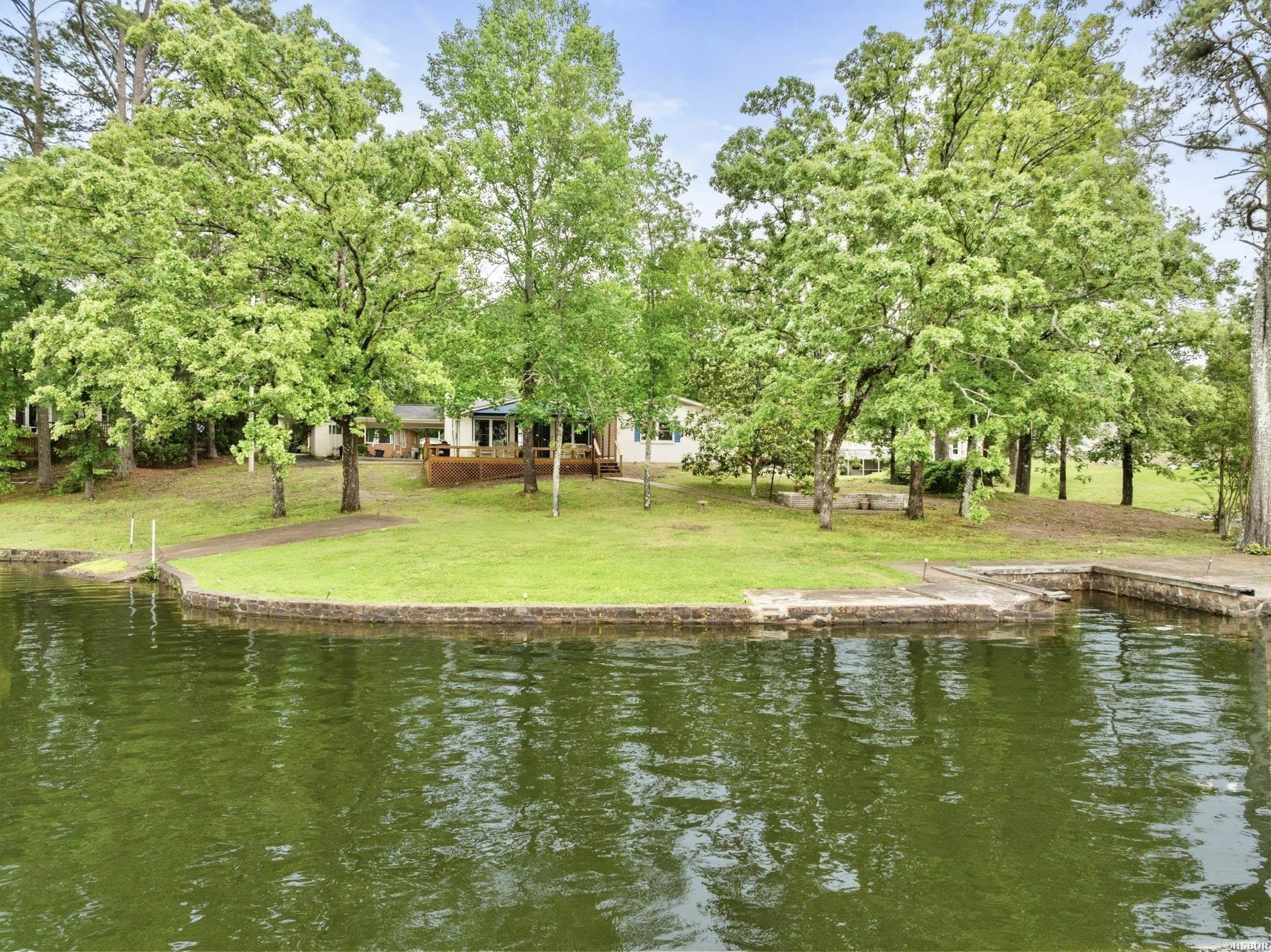 A truly unique opportunity on Lake Hamilton! This beautiful home sits on over half an acre and offers nearly 400 feet of lake frontage, with water wrapping around the property to create breathtaking panoramic views from every angle. The home features 3 bedrooms and 2 bathrooms - all on one level with no steps! The open living area flows seamlessly to a large outdoor deck—perfect for entertaining or relaxing by the lake. Additional highlights include new vinyl plank flooring, woodburning fireplace, newer roof, a two-car carport, two storage buildings for all your lake gear, and even a private boat ramp for easy lake access. Located in the heart of Hot Springs, this property combines convenience with serene lakefront living—just minutes from shopping, dining, and entertainment. Don't miss this rare find with unparalleled lake frontage and endless potential!