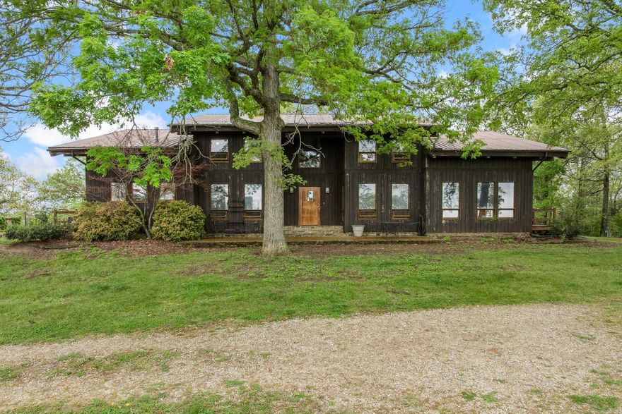 Tucked into the breathtaking Boston Mountains near Huntsville and Osage, this rare Ozark retreat offers unmatched privacy, character, and natural beauty. The stunning 6,434 sq ft contemporary home features 4 bedrooms, 4.5 baths, soaring 26’ ceilings, rich oak and walnut finishes milled from the property, and a dramatic two-story fieldstone fireplace. A finished walk-out basement with workshop connects through a one-of-a-kind stone tunnel to a 3-car garage. A charming 544 sq ft guest cabin with loft, full kitchen, spacious porches, and 2-car carport provides ideal guest or rental space. The 32x50 barn includes electric, 3 stalls, tack room, and pens. The land showcases rolling pasture and timber, fenced and cross-fenced, with 3 ponds, a natural spring, trails, vineyard, and garden space. Shared deep well, mineral rights convey. Just 15 miles to the Buffalo National River—perfect for a private estate, recreation, or investment opportunity.