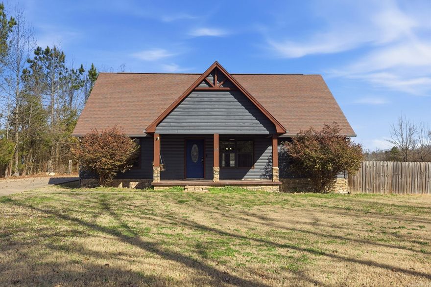 Welcome to this charming rustic-style home nestled on a spacious lot in a peaceful setting. Featuring eye-catching curb appeal, the exterior showcases a warm blend of natural tones, and a welcoming covered front porch—perfect for relaxing and enjoying the surrounding landscape.  Step inside to an inviting open-concept living space highlighted by soaring vaulted ceilings and abundant natural light. The living room is anchored by a beautiful stacked-stone fireplace, adding both warmth and character, while custom wood accents and trim throughout the home enhance its cozy, cabin-inspired feel.  The kitchen seamlessly connects to the main living area and features rich wood cabinetry, granite countertops, stainless steel appliances, and a functional center island—ideal for both everyday living and entertaining.  A standout feature of the home is the custom wood staircase with rustic railing. The polished concrete floors throughout the main level provide durability and a modern touch that complements the home’s natural finishes.  Large windows frame scenic outdoor views, and the generous yard offers plenty of room for recreation, gardening, or future customization.