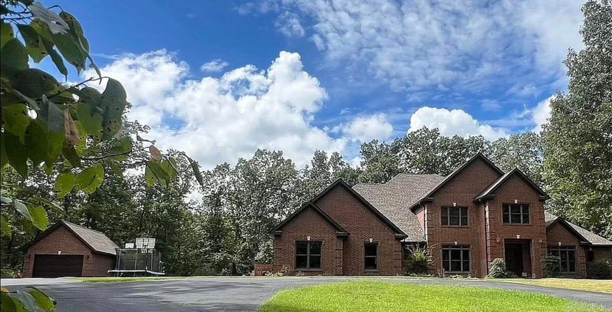 Woodland Hills prestige meets Ozark seclusion in this luxury brick estate on 7 + gated acres bordering protected Ozark Folk Center land—yet still inside Mountain View city limits. A custom solid-oak staircase anchors the two-story foyer and flows into a dramatic 20 × 20 living room where floor-to-ceiling Anderson windows frame tranquil woodland views. Double glass doors reveal a gourmet kitchen with pro-grade appliances, custom cabinetry, and breakfast nook merging into a hearth room with gas fireplace and sightlines to the in-ground pool. The main-level primary suite offers a spa bath and walk-in closet, while a flexible bedroom/office with full bath sits nearby. Hand-scraped Brazilian hardwood and stained concrete floors feature radiant heat that continues into the oversized two-car garage with FEMA-rated safe room. Upstairs: three generous bedrooms (two ensuite) with walk-ins, attic storage, a laundry chute, and a second oak staircase back to the hearth room. Outside, a columned veranda overlooks a 20 × 40 Grecian-style pool surrounded by mature timber, and a 20 × 30 brick garage provides extra parking and storage. Privacy, comfort, and convenience!