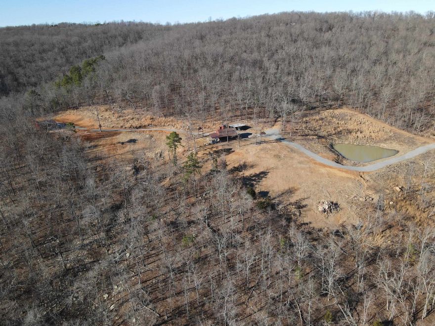 Located at the end of a county road with a gated entrance, this retreat offers privacy and serenity while still being less than a quarter-mile from a paved road and just one-half of a mile from State Highway 16. The first cabin provides approximately 830 sq. ft. of living space, with a cozy 225 sq. ft. covered front porch. It features a metal roof, a drilled well for water, solar power (with nearby electricity available if desired), and a wood cook stove. Inside, you’ll find well-crafted cabinets, a sink, and stairs leading to a loft that overlooks the living area. A bathhouse has been started nearby, and a storage workshop houses the solar components. The second cabin offers 750 sq. ft. of living space and an expansive 512 sq. ft. covered deck with a metal roof. It includes a wood stove, cabinets, and a shower, with a loft that overlooks the living area. Additional amenities include a 20x24 carport, an 8x8 storage building with solar components, and a large 30x40 insulated barn with metal siding and roof. The property overlooks a serene pond with views of the Ozark Mountains in the distance.