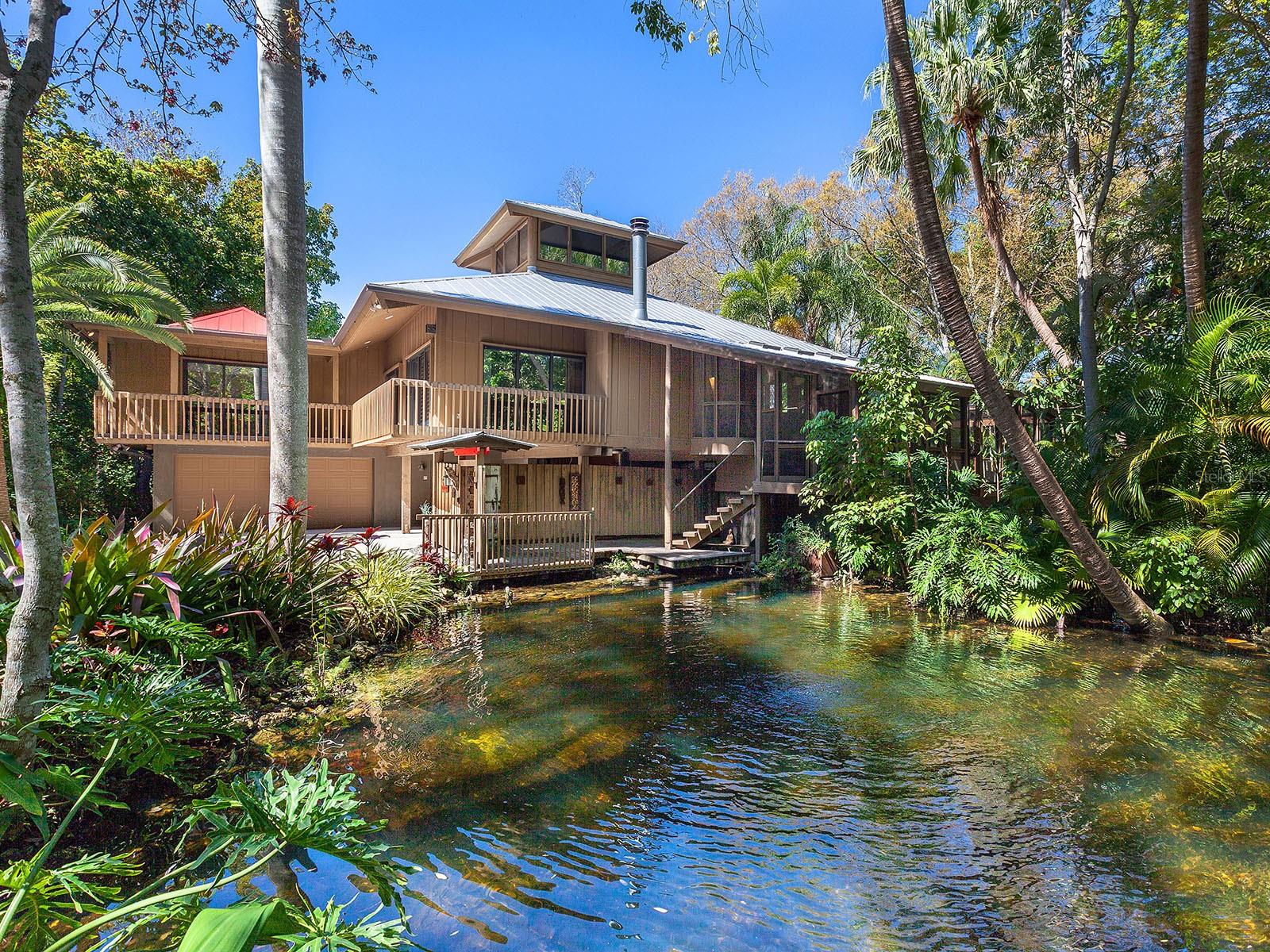 An incredible Sarasota Gem, this East of the trail ‘Treehouse’, designed by Richard G. Allen, a local Sarasota architect known for his innovative and sustainable design ethos, provides a rare opportunity to own an architectural treasure in Arlington Park. This enchanting 3-bedroom, 4-bathroom home with a library/den, offers a truly unique living experience in an exclusive oasis of native Florida ecosystem. Sitting at the end of a private canopied road, surrounded by the lush landscape of protected wetlands, Allen Treehouse and Pond is a masterpiece designed to respect the land and climate using construction techniques and materials local to the Sarasota region. The influence of Frank Lloyd Wright and The Sarasota School of Architecture is felt throughout the home in the way it brings harmony of the surrounding landscape to the indoors and effortlessly combines features of art and nature. 5 minutes from downtown, the home is nestled in a world all your own. As you enter, hand carved Tibetan teak wood gates open to a walkway overlooking a pond view and waterfall feature. The pond has been converted to automatically fill using a well for a seamless integration with the natural water sources and environment. From the ground level, stairs lead to a spacious screened deck and private swimming pool cocooned in the treetops. The home was built and designed to be completely compatible with the Florida climate, blending the organic design, materials and aesthetics with the lush natural environment. The home features broad overhangs, windows and doors situated for airflow, high ceilings, and a remarkable wrap around screened porch. A hand carved door opens into the main living area where you are greeted with maple wood floors, soaring ceilings, exposed beams and expansive windows inviting natural light to dance through the open spaces. Four of the support beams are pine telephone poles hand picked by the architect to encapsulate exposed wood elements. The open concept plan boasts the kitchen, living room, and dining room, with a primary suite and additional guest suite and library all on the main living floor. The kitchen, a central space that ties in the main living areas to the staircase, loft, and two spacious suites, includes upgraded appliances, functional shelving, and enhanced lighting for overall ambiance of the space. On the main level is a visionary library crafted with custom sliding maple doors, beautiful built-ins and shelving placement to blend into the aesthetic of the architectural style. An integral part of the interior design elements, the spiral staircase leads upstairs to an elevated loft with oak wood floors, while the ceiling stretches to an arrangement of clerestory windows that can be opened to the natural light and air. By staircase you can access the ground level from the main floor, which features a private bedroom ensuite with a separate entrance, additional storage and a spacious laundry room. Experience the ultimate convenience with ample parking space, an oversized two car garage, and an electric charging station. This home is an oasis for those seeking a peaceful retreat, while offering a design that is functional and thoughtfully planned to bring the outdoors in, connecting to the beauty of the surrounding landscape. The Allen Treehouse is a sanctuary and piece of Sarasota history for those who appreciate the serenity of nature and the artistry of organic architecture and sustainably minded design.