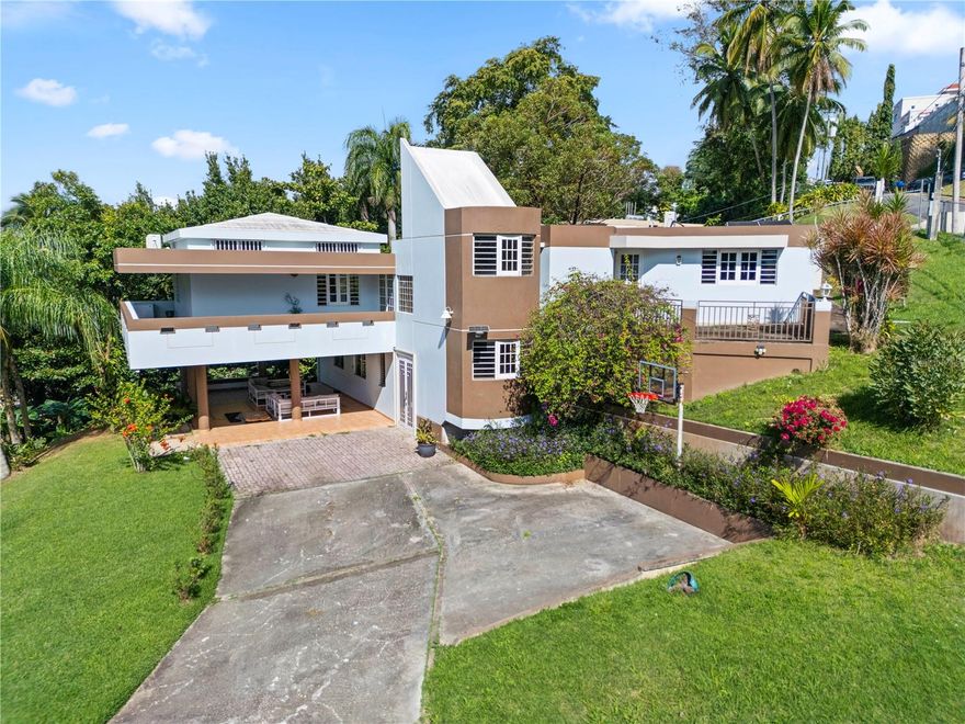 Located within the gated community of El Mirador de San Juan, this distinctive residence offers privacy, space, and a timeless architectural presence on an expansive half-acre lot surrounded by mature tropical landscaping. Avocado and plantain trees frame the property, creating a serene, garden-like setting that feels both established and tranquil.

The home’s architecture blends clean lines with warm, modernist influences—defined by its strong geometric forms, elevated main living areas, and a wrap-around porch that extends the living room outdoors. Covered terraces and shaded outdoor spaces invite year-round indoor-outdoor living, ideal for entertaining or quiet moments surrounded by greenery.

The main residence features 4 bedrooms and 3 bathrooms, with an updated kitchen anchoring the primary living area. On the first level, a private in-law apartment offers exceptional flexibility, complete with one bedroom, one bathroom, and a kitchenette—ideal for guests, extended family, or additional living arrangements.

Designed with efficiency and sustainability in mind, the property is equipped with solar panels and two Tesla Powerwall batteries, providing energy resilience and long-term cost savings.

Conveniently located just 3 minutes from Montehiedra Shopping Center, dining options, and daily conveniences, and only 4 minutes from Highway 52, this home offers a rare combination of privacy, accessibility, and space—set within one of San Juan’s most desirable residential enclaves.

A unique opportunity to own a character-rich home with land, energy independence, and a prime San Juan location.
