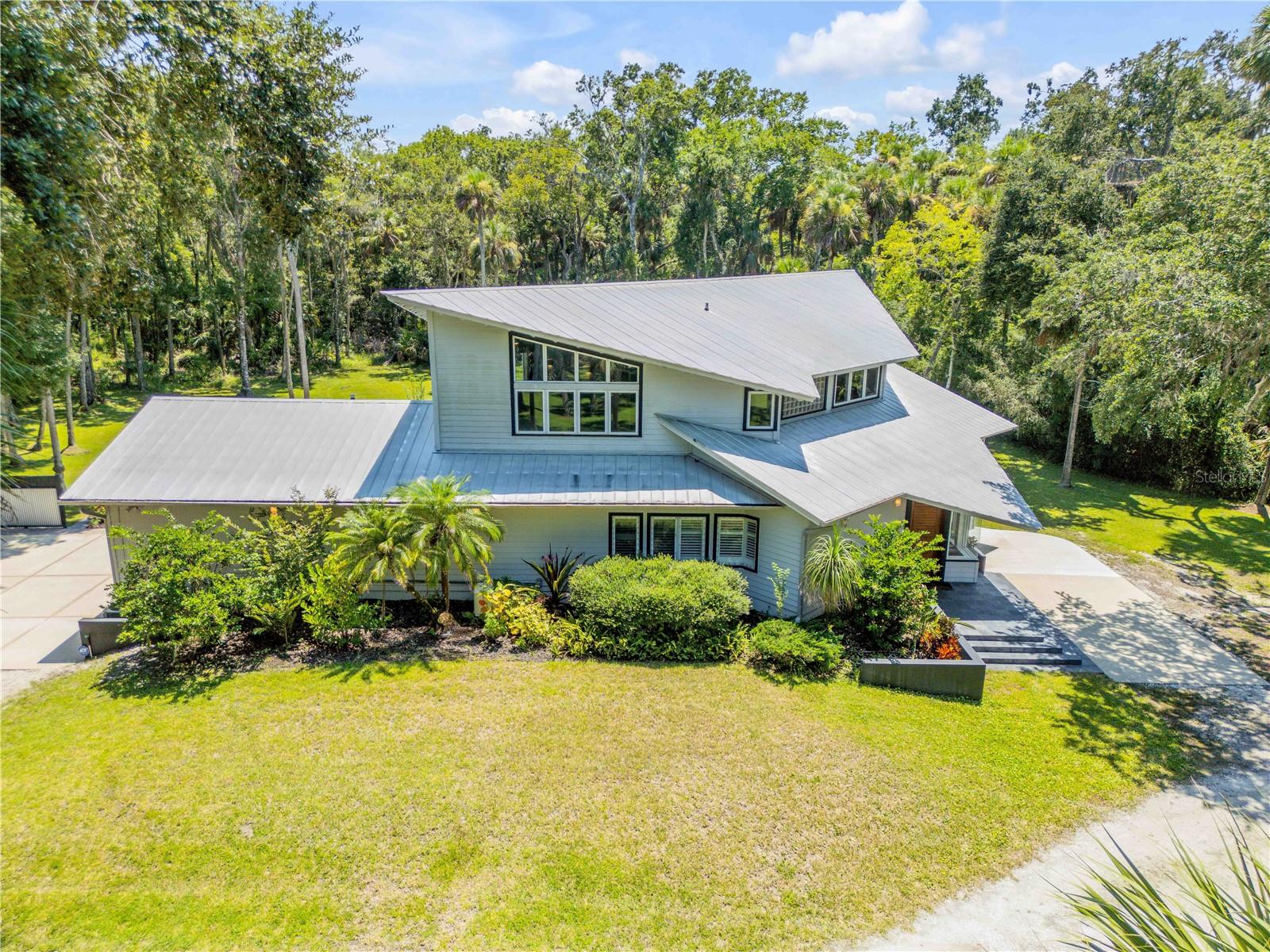 This stunning modern home, designed by renowned architect Chris Frank and built by Hickson Construction, is a rare opportunity to own a piece of architectural artistry in a serene rural setting with the comfort and convenience of city living. Nestled on 3 private acres just minutes from New Smyrna Beach and Port Orange, this custom 3-bedroom, 3-bath residence showcases sleek design, expert craftsmanship, and natural elements that create a harmonious blend of elegance and functionality. From the custom metal gate to the handcrafted front door and built-in bookcase, every detail reflects modern style in a picturesque environment. Inside, exposed beam ceilings and red oak hardwood floors flow seamlessly into natural slate tile in the kitchen, laundry, and bathrooms. The chef’s kitchen features luxury granite countertops and top-tier appliances including a Wolf dual-fuel range, stainless counter-depth refrigerator, and dishwasher. A first-floor bedroom offers privacy and easy access, while a striking open metal staircase leads to a spacious loft with soaring ceilings and a wall of windows—perfect for a home office or additional living space. Upstairs, the owner’s suite is a true retreat with an expansive bath featuring a soaking tub, separate walk-in shower, and walk-in dressing closet with built-in drawers. The property includes an oversized attached two-car garage and a detached, air-conditioned single-car garage that currently serves as an ideal office, studio, or gym with its sliding glass door and climate control but can easily be converted back to a traditional garage if desired. Featuring double-paned, hurricane-rated windows and doors, solid wood interior doors, a 22kw Generac whole-house generator, on-demand propane water heater, and two updated HVAC systems, this home is designed for both comfort and resilience. Outdoors, a canopy of oaks surrounds the custom paver patio with stone firepit, gazebo, and outdoor bath, leading to a screened back porch overlooking a manicured lawn—perfect for entertaining or quiet reflection. The property also provides space to add an additional building if desired, with the owner having started the process of constructing a 42x60 metal building. Ideally located near Canter Down Farms, The Preserve at Turnbull Bay Golf Course, and Doris Leeper Spruce Creek Nature Preserve, this property offers the perfect balance of nature and convenience.