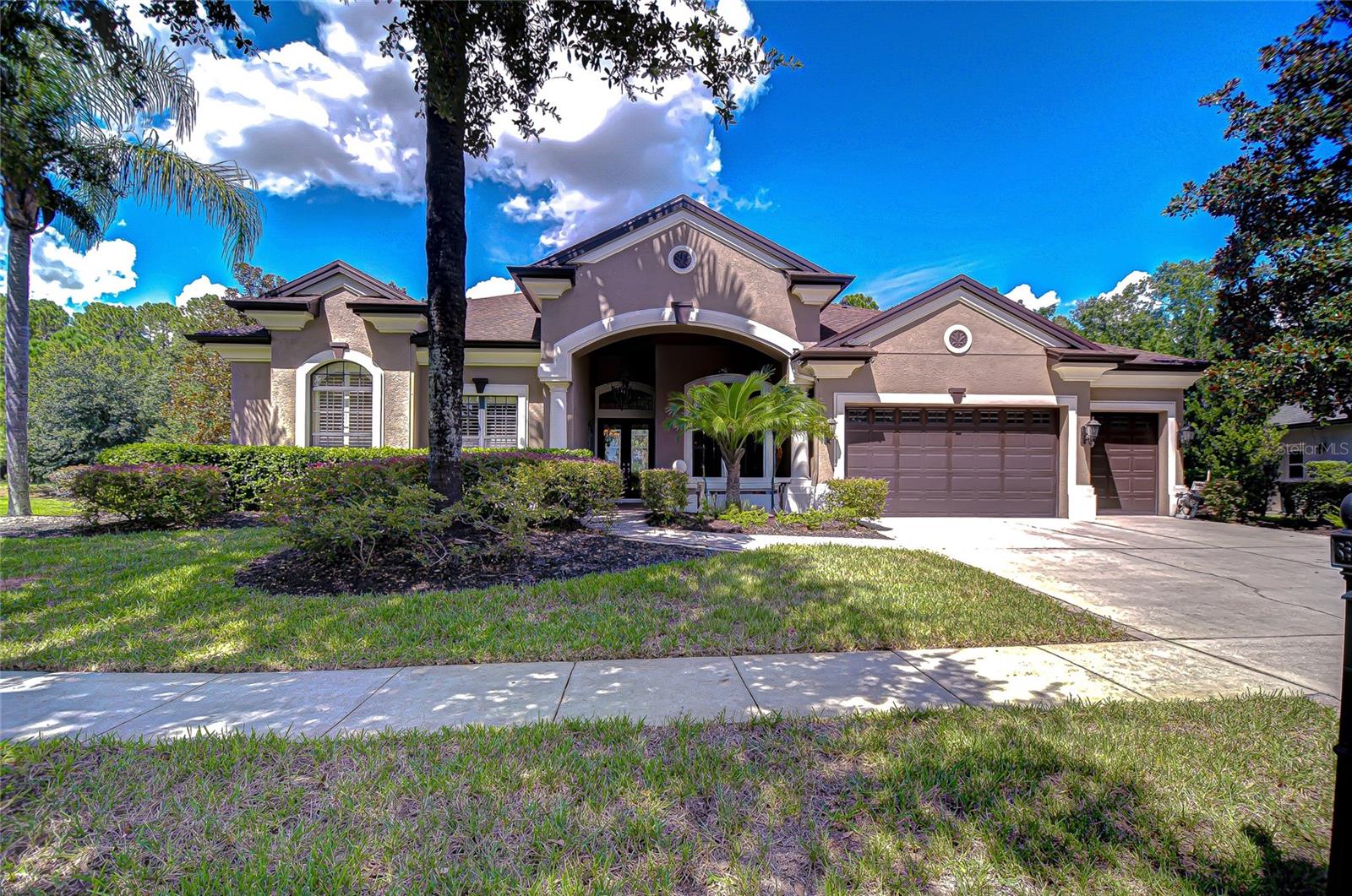 Stately executive residence on a picturesque wooded cul-de-sac with a tranquil side pond and back stream, this Arthur Rutenberg St. Augustine V model is perfectly situated in the exclusive, highly sought-after GATED waterfront community of Pasco Sunset Lakes.
From the moment you arrive, the portico-covered entrance framed by tall pillars and a welcoming front porch accented with half-pillars sets the tone. NEW Roof 2024! HVAC 2021! Glass double front doors invite you inside, where timeless architectural details and traditional charm blend seamlessly with an open-concept design. This expansive 1-story home boasts 3,609 sq ft with 4-Bedrms, 3.5 Baths, a 3-Car garage. Soaring ceilings, crown molding, Plantation shutters, archways, columns, rounded corners, and a desirable 3-way split-bedroom layout provide ultimate privacy for families and guests.
The gleaming ceramic tile floors flow throughout the main living areas, beginning with a dramatic foyer. Here, French double doors open to a large office with rich hardwood floors, while a stylish guest bath with pedestal sink and mantle sits just across. A pony wall with a decorative column separates the foyer from the formal dining room, enhanced by a tall, arched window that fills the space with natural light.
At the heart of the home, the chef’s kitchen impresses with 42-inch solid wood cabinetry topped with crown and display space above. A tile backsplash with decorative inlays complements the granite counters, while an extended hi-top breakfast bar and center island create plenty of space for cooking and gathering. Premium appliances include a Professional Wolf natural gas cooktop with vent hood, GE Monogram double ovens with convection and microwave, built-in refrigerator, and Bosch dishwasher. A separate dining area with bay windows overlooks the sparkling pool.
The living and family rooms, each adorned with sliding Plantation shutter panels, open wide with pocketing sliders to reveal the screened lanai, paver deck, and heated pool with a spill-over sheer descent—a private outdoor oasis that blurs the line between indoor and outdoor living. The family room features a tray ceiling, while the living room showcases a striking coffered ceiling, fireplace with mantle, and custom built-ins with glass shelving. This thoughtful design creates the perfect balance of elegance and warmth for everyday living and entertaining.
The opulent primary bedroom retreat with rich designer carpet and stunning tray ceiling has a sitting area with bay windows crown molding and French door access to the pool. Two separate walk-in closets contain pull-outs, shelves, drawers, bins, and more. The luxurious ensuite bath features a tray ceiling, crown molding, dual granite topped vanities, a make-up area, a deep soaking Jacuzzi tub with spa jets framed by columns and an arched window, a walk-in tiled shower with Listello accent, and private water closet.
Two additional bedrooms share a Jack and Jill bath, while the fourth bedroom/bonus/flex space features convenient access to a full pool bath and French door leading to the picturesque backyard—an inviting setting where you can relax with peaceful views and the chance to spot local wildlife.
Residents enjoy exclusive access to a private beach club, boat ramp, 42-acre ski-lake, playground, more. Minutes from major highways, shopping, dining, and top-rated schools. Don’t miss this rare opportunity—schedule your private showing today and make this dream home yours!