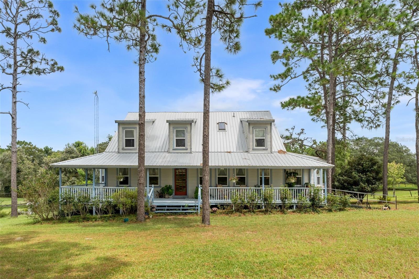 This storybook-style farmhouse offers timeless charm, peaceful acreage, and equestrian-ready amenities—just minutes from Ocala, the Horse Capital of the World. Set on 6+ quiet acres along a scenic limestone road, the property is conveniently located near trailheads to Goethe State Forest, and just a short drive from WEC, HITS, Black Prong, and all the best of Marion County horse country.
With nearly 6,000 sq ft under roof, the home includes 2,726 sq ft of heated living space plus a 1,793 sq ft walkout basement with a private entrance that can easily be finished—perfect for a guest suite, in-law apartment, or income opportunity. The great room welcomes you with a soaring 27-foot cathedral ceiling, a gas stone fireplace, and a hand-painted tree mural that brings a whimsical touch to the space. The kitchen features solid cherry cabinetry and countertops, with both a breakfast nook and formal dining room nearby. The primary suite connects to a private bath and a dedicated office, while upstairs includes two guest rooms, a full bath, a bonus room, and hidden storage tucked under the eaves.
Step outside to a nearly full wraparound porch, where sunrises and sunsets paint the sky and life naturally slows down. It’s the perfect spot to sip coffee, watch the kids play, or simply breathe.
The 6 stall center isle barn includes a tack room and loft area. Two RV hookups offer added flexibility for guests or seasonal visitors. A newer roof and A/C unit provide peace of mind, while the location offers unmatched access to world-class equestrian venues, trail riding, and small-town charm.
Whether you’re growing your family, starting your dream farm, or simply looking for room to roam, this property offers the lifestyle you’ve been searching for.