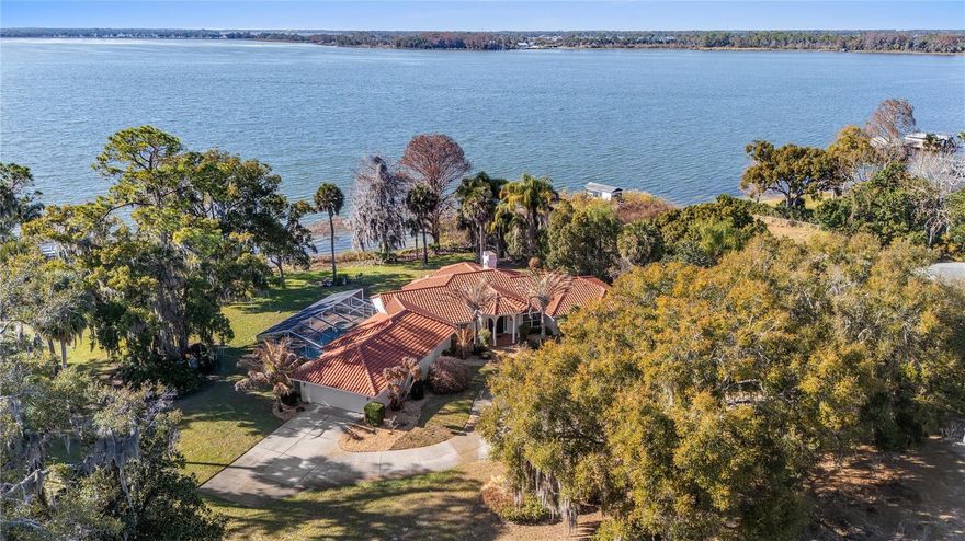 Envision the arrival of your private lakefront estate on the storied shores of Lake Beauclair with direct access to Lake Dora, the crown jewel of the Harris Chain of Lakes, a vast and celebrated waterway offering endless miles of lock and river cruising for days defined by leisure and adventure. Life on the lake unfolds at a gentler, more inspired pace as mornings begin with coffee on the dock while mist rises from the water, afternoons invite boating, paddleboarding and fishing along scenic shorelines, and evenings conclude with unforgettable sunset cruises beneath painted skies. Just moments away, the charm of Mount Dora enriches the lifestyle even further. Known for its storybook streets, vibrant arts scene and year-round calendar of festivals, residents enjoy boutique shopping, waterfront dining, antique markets, boat shows, food and wine celebrations, and lively community gatherings that make every season feel like an occasion. Across a remarkable eight-acre parcel, 6136 Dora Drive commands hundreds of feet of pristine shoreline, ideally oriented to the west for breathtaking sunset vistas that paint the water in gold each evening. This extraordinary canvas invites limitless possibilities such as design a grand equestrian compound, cultivate a private orchard, create an inspired guest residence with an expansive collector’s garage or explore opportunity to thoughtfully subdivide. Within a canopy of mature trees, a sprawling single-story residence spans approximately 4,000 square feet, offering three bedrooms, two and a half baths, walls of windows, a screened pool and spa, and a newer tile roof. Completing this rare offering is an enclosed boathouse with private doors, an increasingly scarce luxury along the chain. With no HOA restrictions and boundless space to create, this is a legacy lakefront setting where vision meets lifestyle, a place where lake days, sunset evenings and timeless charm of Mount Dora define life at its most exceptional.