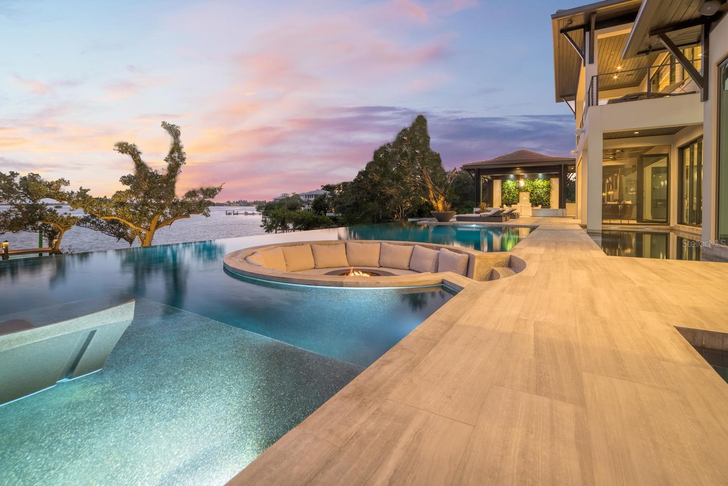 Relax and take in the positive energy of Lotus-style living in this one-of-a-kind Balinese-inspired resort compound on north end of the world-famous Siesta Key. Nothing is needed to begin your Zen journey as the property comes completely furnished down to dishes, silverware and a Buddha living wall. The coexistence of indoor and outdoor space blends over 8,000 square feet of indoor space with thousands of square feet of patio and pool area. An outdoor oasis offering an infinity-edge pool with a swim-up bar, a sunken fire pit lounge within the pool, a spa, an outdoor kitchen, a dining pavilion with a living plant wall and an open-air living room with a fireplace. A koi pond with stunning built-in footbridges stretches nearly the entire length of the home, enhancing the serene ambiance of the outdoor space. Spanning over an acre with more than 700 feet of waterfront, the property is a masterpiece of luxury, designed for effortless relaxed living or island-style entertaining. A custom-designed driveway welcomes you to a breathtaking entrance, where waterfalls and fire features set the tone for the experience that awaits inside. Expansive walls of glass, seamless indoor-outdoor transitions, and rich wood and stone flooring throughout create a striking, cohesive aesthetic. The thoughtfully designed interior boasts multiple living spaces, including a game room, home theater, fitness room, meditation space and an impressive 8-plus-car garage. For the waterfront enthusiast, a well-equipped boat dock, a separate waterfront garage, and a paddleboard/canoe launch provide unparalleled access to pristine waters. This exceptional residence also features a newer seawall, full-house generator, Crestron home automation, a three-stop elevator and a whole-house reverse-osmosis water filtration system, ensuring convenience and peace of mind. An island paradise, this estate offers unmatched privacy while remaining just moments from Sarasota’s vibrant shopping, dining and world-renowned cultural attractions. Don’t miss this rare opportunity to own a move-in-ready masterpiece on Siesta Key – home to the #1 beach in the USA (Tripadvisor).