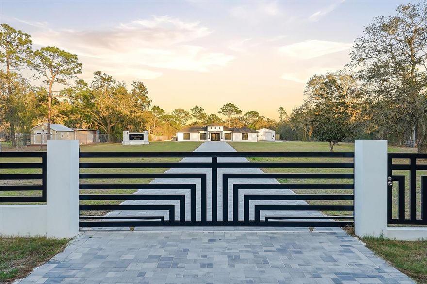 Welcome to this fully custom masterpiece in Spring Hill, where modern luxury meets privacy and functionality on 2.5 acres of beautifully cleared land. This exceptional residence offers 3 bedrooms and 2.5 bathrooms, thoughtfully designed with elevated finishes and impeccable craftsmanship throughout. From the moment you arrive, the attention to detail is undeniable. The home features a spacious two car attached garage with sleek epoxy flooring. Inside, a dramatic accent wall in the living room creates a striking focal point, perfectly complementing the expansive open concept floor plan designed for refined entertaining. The chef inspired kitchen is a true showpiece, appointed with high end acrylic cabinetry, soft close drawers, premium appliances, and an oversized island ideal for gathering. Modern illuminated bathroom mirrors, designer light fixtures, and high ceiling and luxury ceiling fans enhance the contemporary aesthetic, while abundant natural light pours into every space. Throughout the home, 96” solid core Euro style interior doors add an elegant architectural touch. The owner’s suite serves as a private retreat, featuring an expansive walk-in closet and a spa inspired bath with a must see oversized walk in shower, designer tilework, and elevated finishes. The additional bedrooms are generously sized, and the half bath is perfectly positioned for guests. Built with quality and efficiency in mind, the property includes a 2024 roof, AC system, septic and well system with Clack filtration, and energy efficient windows, offering peace of mind and modern sustainability. Beyond the main residence, the estate continues to impress with a fully detached metal building, ideal for storage, business, or hobby use. This impressive structure includes a commercial grade air compressor and a BendPak two-post lift, a rare and valuable addition. The exterior is equally exceptional, featuring custom concrete and aluminum fencing with a main electric gate, two additional vehicle gates, and a walk in entry for privacy and security. Professionally installed pavers create elegant outdoor living spaces, highlighted by a covered outdoor kitchen complete with double convection oven combos, two grills, a griddle, a modern fire pit with seating areas, a central island, and a full contemporary outdoor bathroom, perfectly designed for unforgettable gatherings and elevated entertaining. Ideally located near major roadways, fine dining, and shopping, this extraordinary property offers the perfect balance of luxury, space, and convenience.This is more than a home ,it is a statement of lifestyle, craftsmanship, and elevated living.