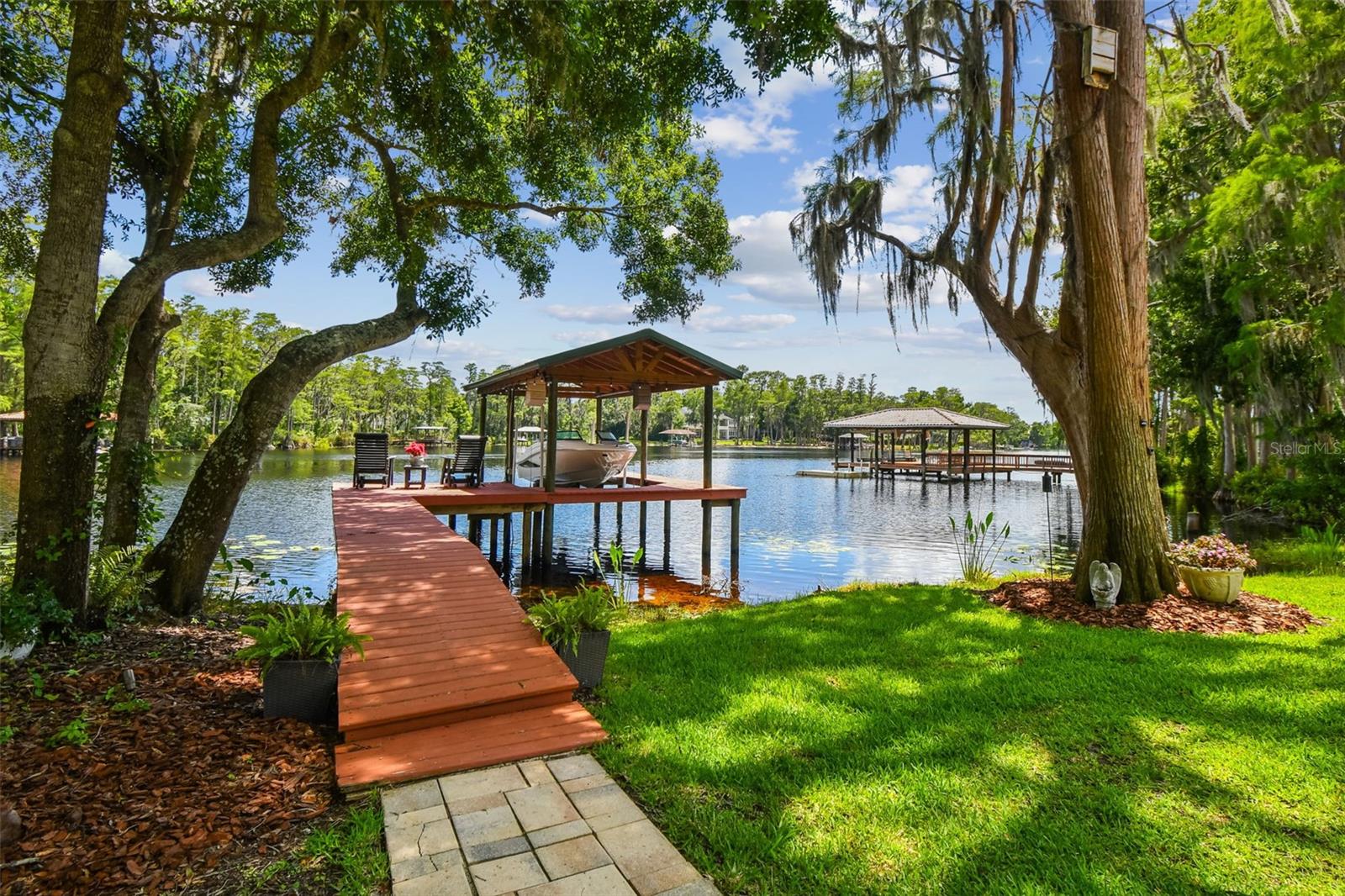 Experience LAKEFRONT LIVING at its finest in an exclusive, 10-home GATED enclave on a private 0.85-acre SKI-LAKE cul-de-sac lot in coveted Pretty Lake Estates on LAKE PRETTY. This one-story waterfront home offers 4 bedrooms and 3 1/2 bath, with stunning lake views from every room. A long, private driveway leads to a double-door entry flanked by a tranquil koi pond and generous parking.
Soaring ceilings and slate flooring transition seamlessly into hand-scraped Brazilian cherry wood, framing the living room’s WOOD-BURNING FIREPLACE and showcasing sweeping panoramic views. The CHEF’S KITCHEN impresses with dual islands topped in striking EXOTIC GRANITE, floor-to-ceiling wood cabinetry, TRAVERTINE flooring, and select newer appliances. It opens effortlessly into a SECOND FAMILY ROOM, perfect for everyday comfort or entertaining guests.
The primary suite is a peaceful retreat with dual walk-in closets and SWEEPING LAKE VIEWS. Its spa-like bath features marble floors, a rain shower with built-in seat, and floor-to-ceiling marble tile. Two guest bedrooms share a JACK-AND-JILL bath and a glass-enclosed shower that also serves as the POOL BATH.
Step outside to your PRIVATE PARADISE, a covered patio with skylights and ceiling fans shelters the OUTDOOR KITCHEN w/full wet bar and grilling station clad in stacked stone and dark granite, complete with sink, storage, grill, and draft-beer tap. Beyond, an oversized, SALTWATER MARCITE POOL rests on a flagstone deck, surrounded by paver patios, lush landscaping, and tasteful outdoor lighting beneath mature oaks. With over 140 feet of LAKE FRONTAGE, you’ll enjoy a private BOAT DOCK WITH LIFT; a 2012 Sea Ray 190 Sport Boat is available separately for effortless aquatic adventures. Enjoy the SERENE BEAUTY OF NATURE as you watch and listen to a symphony of birdsong from a variety of birds right in your own backyard.
NO FLOOD ZONE! Upgrades include a 300-ft deep WELL, enhanced pool pump and water-conditioning system, refreshed landscaping with upgraded lighting, dual water heaters, a NEW PLATINUM-PROTECTED ROOF, and an updated electrical panel. HURRICANE-RATED WINDOWS AND DOORS provide added peace of mind. PRETTY LAKE ESTATES is located just a few miles from scenic trails, bike paths, and dog parks, and with the Suncoast/Veterans Expressway offering quick access to Downtown Tampa, the airport, shops, dining, and top-rated STEINBRENNER HIGH, this Odessa retreat delivers unparalleled lakefront elegance and everyday functionality. Schedule your private showing today!