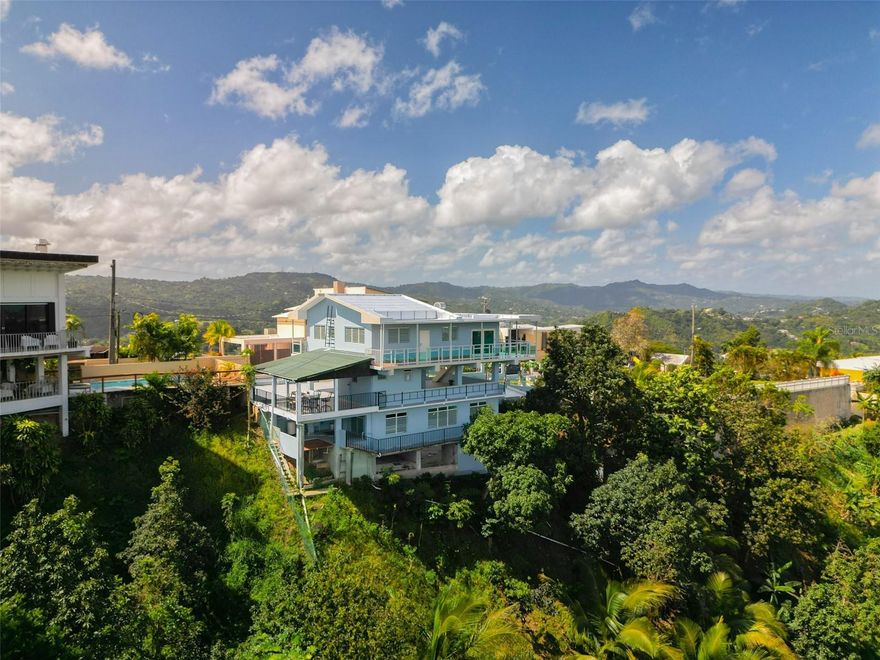 HOLLYWOOD HILLS — JUST 10 MINUTES FROM THE SAN JUAN METROPOLITAN AREA — Private hillside estate with extraordinary 180-degree panoramic views spanning the south, west, and north horizons. This custom-built three-level residence offers a rare combination of privacy, versatility, and breathtaking natural scenery. The main upper-level residence features approximately 1,470 square feet of living space with a 3-bedroom, 2-bath layout. Expansive glass openings and a wraparound balcony capture sweeping mountain and sky views while allowing abundant natural light and seamless indoor–outdoor living. The lower level includes a fully independent 2-bedroom, 1-bath guest residence with a private terrace, ideal for extended family, guest accommodations, executive retreat use, or potential income-producing opportunities. Recently renovated with hurricane-resistant doors and windows, providing durability and peace of mind. Situated on an expansive 4,290-square-meter hillside lot, the property offers exceptional privacy and a unique topography that enhances its panoramic vistas. The land also presents opportunities for future expansion, terraced gardens, or customized outdoor living spaces. 
Additional property features include a spacious open-air terrace and lounge area, separate storage / tool structure, covered carport for up to four vehicles, private water well system with approx. 7,000 gallons of reserve capacity, etc.  — Offered at appraised value and open to qualified offers. For additional information or to schedule a private showing, call today — ¡Llame hoy!