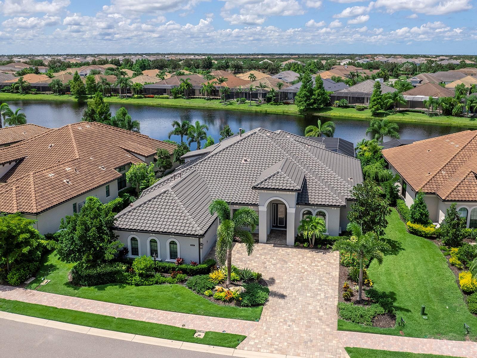 On a tranquil lakefront home site in Country Club East, built by Cardel Homes, this Dolcetto 4 floor plan offers over 3,300 square feet of refined living, where a natural color palette and wood-look tile floors set a tranquil tone. Thoughtful upgrades—custom ceilings, crown molding, designer lighting and detailed trim—blend craftsmanship with comfort throughout. A paver driveway and lush landscaping introduce the home, with no neighbors across the street—just a tranquil walking path and gazebo. A stunning glass front door, framed by side windows and a transom arch, opens to a foyer with a wallpaper-accented niche, tray ceiling, crown molding and coffered details—all set above elegant wood-look tile floors that continue throughout the main living areas. The great room offers a gas fireplace with a carved stone mantle, custom coffered ceilings, crown molding and transom windows above pocketing sliders with electric shades, all framing peaceful lake views. The adjacent kitchen showcases custom cabinetry, quartz countertops, a stainless-steel farm sink, under-cabinet lighting and top-tier KitchenAid appliances—including a six-burner gas cooktop with a vent hood. A spacious walk-in pantry with wood shelving and convenient pullout drawers offers abundant storage. Centered by a large island with pendant lighting, the space flows seamlessly into the open dining area, surrounded by designer windows and detailed wainscoting. Designed for entertaining, the outdoor living area includes a covered summer kitchen with polycarbonate cabinetry, granite countertops, a built-in grill with custom-trimmed vent, remote-controlled screens, and architectural columns that divide lounge and dining zones. A lap pool and spa with blue and teal tile and a water spillover feature complete the lakefront retreat. A custom archway leads to the private guest wing, featuring two spacious bedrooms with walk-in closets, large windows, plush carpet, and a shared Jack and Jill bath with dual vanities, a walk-in shower and subway tile. A separate pool bath, accessible from the bonus room, offers a glass-enclosed shower, designer wallpaper and a solid surface vanity. The primary suite is privately situated and designed for serenity. The bedroom features plush carpet, custom-trimmed coffered ceilings and numerous windows for natural light. The luxurious bath includes a barrel-style ceiling with a chandelier, crown molding, dual quartz-topped vanities, LED-lit mirrors, a free-standing soaking tub, large glass-enclosed walk-in shower with a bench, tile floors and walls, a linen closet and two custom walk-in closets. The office/den features glass French doors, a coffered ceiling with a designer chandelier, a custom accent wall, and a built-in illuminated bookshelf. A nearby bonus room, accessed through a trimmed archway, offers flexible use with crown molding, wood-look tile and plantation-shuttered windows. Additional highlights include a valet drop zone with built-in workspace and storage, an oversized side-entry garage with custom shelving, ceiling racks, plantation-shuttered windows, a Kohler whole-house generator, dual HVAC systems, a tankless water heater and a central vacuum. The laundry room offers cabinetry, folding space, a hanging area, crown molding and a plantation-shuttered window. Country Club East residents enjoy a private clubhouse with fitness center, resort-style pools and proximity to Waterside Place, LWR Main Street and The Mall at UTC.