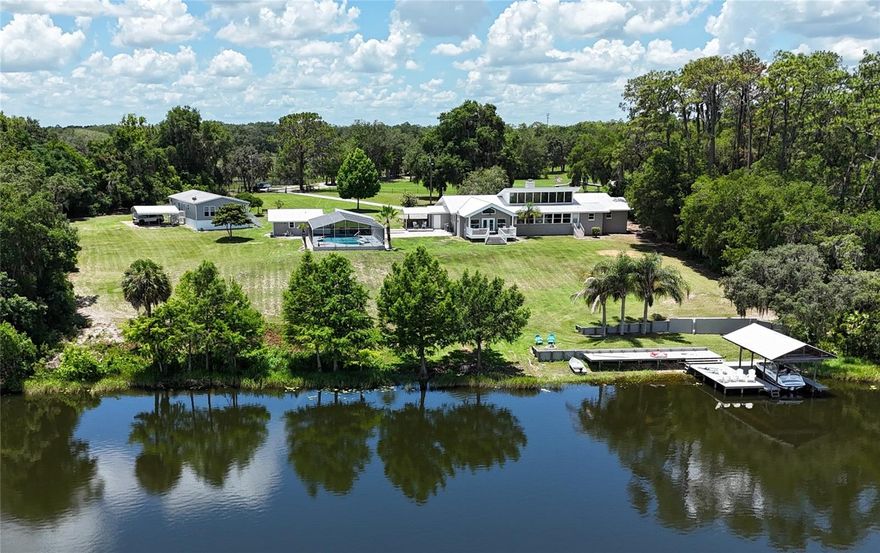 Premier Lakefront Living — Where Elegance Meets Tranquility
Experience the perfect blend of elegance and rural charm with this stunning lakefront estate featuring over 500 feet of private, spring-fed shoreline on Lake Fay. Situated on 14.75± acres and zoned A1, the property is ideal for equestrian or cattle use. The fully remodeled main residence includes a custom addition designed to capture natural light and panoramic views from sunrise to sunset. The spacious living area features a stone wood-burning fireplace, and the open-concept kitchen flows seamlessly into a large dining area, perfect for entertaining. The luxurious owner’s suite offers a gas fireplace, vaulted ceilings, French doors, expansive windows with private deck access, a coffee and wine bar, an elegant marble bathroom with dual vanities and a spa-style shower, and a 14' x 14' walk-in closet. The home also includes a second ensuite, plus two additional generously sized bedrooms, each with walk-in closets and thoughtful separation for privacy. Outdoor amenities include a screened 9-foot-deep pool, a cabana, an outdoor kitchen, and incredible pool-to-lake infinity views. A private dock with sun deck and boat lift enhances lakeside living. Additional features include a newer metal roof, three A/C zones, water softeners, Icynene insulation, a separate 2BR/2BA guest house with its own well and septic, a 60' x 40' equipment barn, an open-air two-stall barn, and fully fenced and gated grounds. This exceptional property is a rare opportunity for those seeking luxury, privacy, and space, just 20 minutes from The Villages.