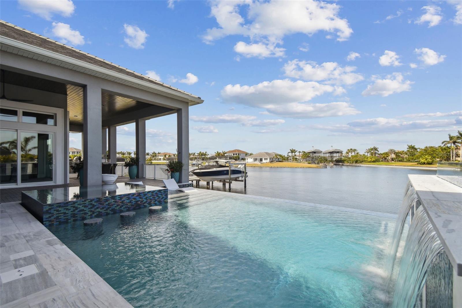 Live the dream in this one-story, fully customized waterfront estate located in the prestigious gated Mirabay community. Located on a private lagoon with direct Bay to Gulf access, this home offers 100 feet of water frontage, dock with Trex decking, and a maintenance free 16K-lb remote-controlled boat lift. Outdoor living takes center stage with a 1,250 sq ft covered patio, Synergy wood ceilings, and a new outdoor kitchen built to impress. Outfitted with three premium grills including a 40" natural gas and custom Gaucho, ice maker, sink, fridge, and stone countertops and backsplash, it’s the ultimate space for entertaining. The resort-style heated pool and spa is truly breathtaking—featuring a dual infinity edge, an oversized champagne spa, sun shelf, swim-up bar stools, fire and waterfall features, and color-changing lighting throughout. Surrounded by glass tile, leather marble coping, and tropical landscaping, it’s all designed for low maintenance with fully automated systems. This home offers water views from every living space and the owner’s suite, making it the perfect backdrop for both entertaining and peaceful relaxation. The Dining Room features a 15' step up tray ceiling with crown molding and is adjacent to the Living Room with an equally stunning 15-ft coffered tray ceiling, dramatic aged brick accent wall, a three-sided electric heated fireplace, and a built-in bar complete with ice maker, glazed subway tile backsplash, 42” upper glass cabinets, and LED accent lighting—an entertainer’s dream. The main living area stuns with 15-ft coffered ceilings with crown molding and custom wood inlay, electric heated fireplace and pocketing sliders, accented by custom wooden barn doors, that open fully to merge indoor and outdoor living, revealing panoramic water views and a true resort-style backyard. At the heart of the home, the chef’s kitchen combines beauty and function with Monogram Professional appliances featuring three ovens, warming tray, pot filler, dual backsplashes in glazed subway tile and ledger stone, rich 12-inch ceiling beams, and designer lighting. The owner’s suite is a luxurious retreat, boasting 13-ft tray ceilings with 12-inch ceiling beams, automated blackout blinds, and 10-ft custom barn doors that open to an expansive custom walk-in closet. The ensuite bath offers a spa-like escape with 12-ft ceilings, an aromatherapy dual shower system with rain head, freestanding Kohler tub, floor-to-ceiling designer tile, glass shelving, and backlit dehumidifying mirrors. Each of the four bedrooms includes its own ensuite bath, custom walk-in closets, and elevated finishes, while a dedicated office/media room with double doors adds privacy and flexibility. Every inch of this home has been thoughtfully upgraded with timeless finishes and luxury in mind including a fully customized laundry room, hallway coffee bar and 141 bottle wine cellar. Entire home features porcelain tile flooring, 7 1/4" baseboards, Plantation shutters, Cambria Quartz countertops, designer lighting and hardware, recessed LED lighting, hurricane impact doors, windows and sliders ~ SO MANY UPGRADES ~ please refer to the FEATURES & UPGRADES list in the attachments. Live every day like you're on vacation with resort style amenities including clubhouse, cafe, 2 heated pools, pickleball, tennis, basketball, sand volleyball, fitness center, kayaks, or enjoy biking endless miles of scenic nature trails. This truly is the finest in waterfront living.