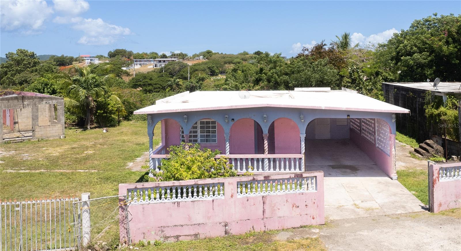 Tucked into the Bravos neighborhood and just a short walk from La Chata beach, this property offers an intriguing mix of history, space, and opportunity. Once part of a sugar mill, the main house stands solid with character and good bones — ready for someone with vision to bring it back to life. It features a bright and spacious great room, four comfortably sized bedrooms, one full bath, and a full functionally sized kitchen that’s ready for remodeling. A front balcony provides a quiet spot to enjoy morning or afternoon coffee, and the covered carport fits two vehicles with ease.
Beyond the main home, there’s a second unfinished structure set up as a 3-bedroom, 1-bath layout with its own living room, kitchen, and dedicated parking area. It will need a roof but offers a strong foundation for guest use, rental potential, or a multi-family setup.
The lot itself is over a third of an acre, with two gated entrances and plenty of space to add a pool, create a garden, or build out outdoor living areas. It’s just a short walk to La Chata beach and a three minute drive from town while still feeling tucked away in a walkable, well-known neighborhood. This is a solid property in a great location with history, substance, and room to grow.