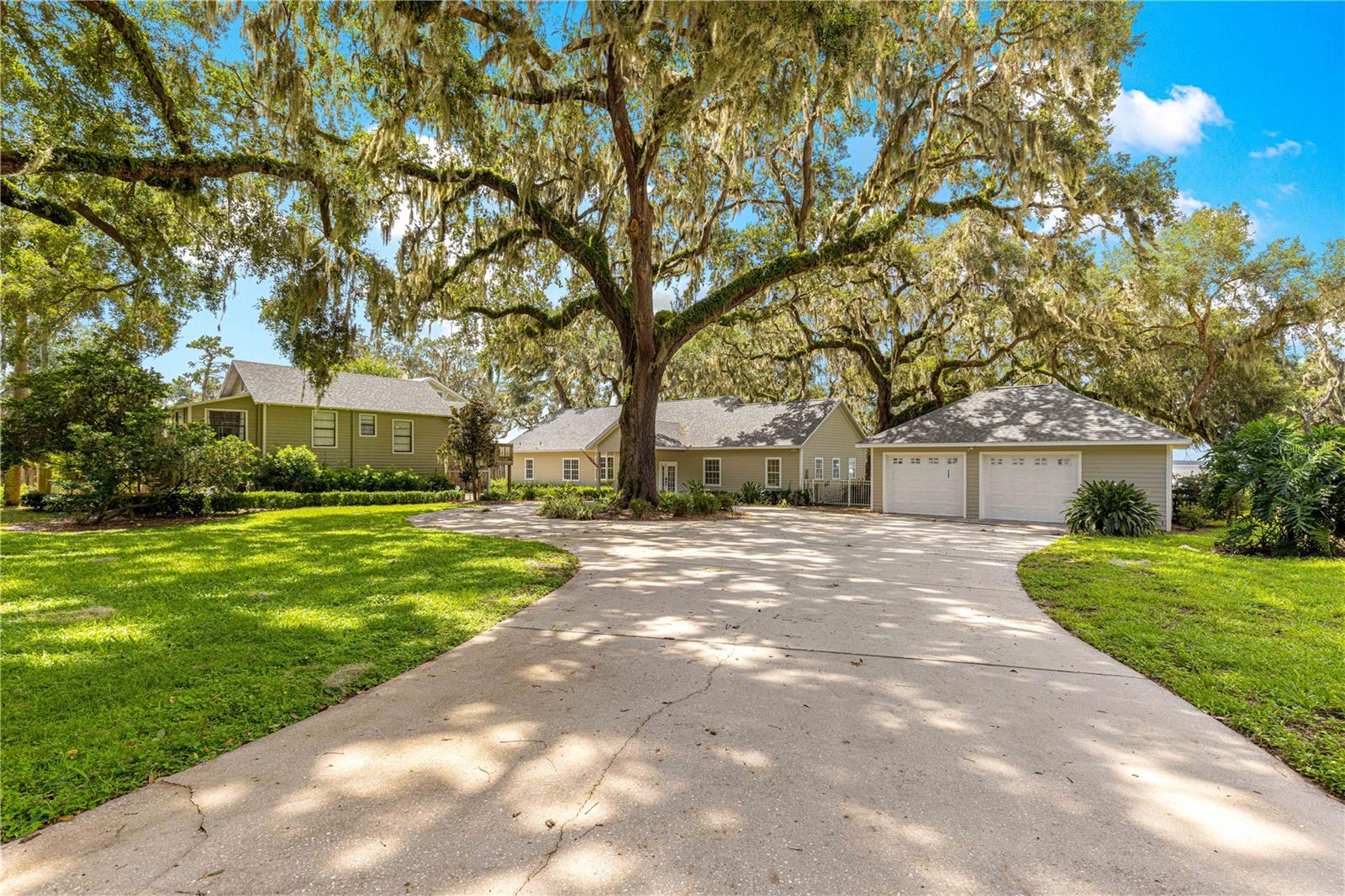 This exceptional luxury estate on the highly sought-after shores of Lake Weir offers an unparalleled lakefront lifestyle with approximately 168 feet of pristine sand beach. Set on a beautifully landscaped 1.79-acre parcel, this expansive property features a thoughtfully designed split-plan main home with over 2,800 square feet of living space on the first floor, plus two separate upstairs areas offering incredible flexibility. A detached 1-bedroom, 1-bath guest cottage with full kitchen, living room, and storage underneath adds charm and versatility to this remarkable waterfront retreat.
Inside the main residence, a long central hallway stretches from front to rear, creating a distinctive “dog run” layout and leading to a huge, enclosed back porch overlooking the shaded backyard and sparkling lake. The oversized living room, with soaring ceilings and beautiful hardwood flooring, flows seamlessly into a spacious kitchen offering abundant cabinetry, generous surface space, and a dedicated butler’s pantry/storage area.
The luxurious downstairs primary suite includes a lower-level seating area and a glamour bath. A private bonus room above the suite, accessible via the master suite, features a Juliette balcony that overlooks the open living, dining, and kitchen areas below. Off the main living space, a full set of stairs leads to an upper landing with a large bedroom, closet, half bath, and lake views, as well as access to the mechanical room housing the home’s HVAC system and water heater. Two additional generously sized bedrooms on the main floor share a spacious Jack and Jill bathroom, while a half bath with shower is conveniently located next to the laundry room, which includes a sink, washer/dryer hookups, and pocket doors for optimal space efficiency.
The outdoor living areas are equally impressive, with gorgeous shade trees, a large front yard, and a long gated driveway that leads to a paved circular drive. A second gated drive offers direct backyard access—perfect for boats and lake equipment. The expanded dock system features a covered boathouse with lift and a water slide, ideal for accessing and enjoying popular and gorgeous Lake Weir, from boating and water sports to breathtaking sunsets.
Additional highlights include a detached two-car garage with two standard garage doors and a third oversized door that opens toward the lake, Hardiboard siding on all structures, underground electric, and a central vacuum system (condition unknown). With its bright, airy layout, versatile floor plan, and massive potential, this rare lakefront gem offers a once-in-a-lifetime opportunity to create one of Lake Weir’s most desirable estates. A few final cosmetic updates and personal touches will elevate this exceptional property to its full potential.