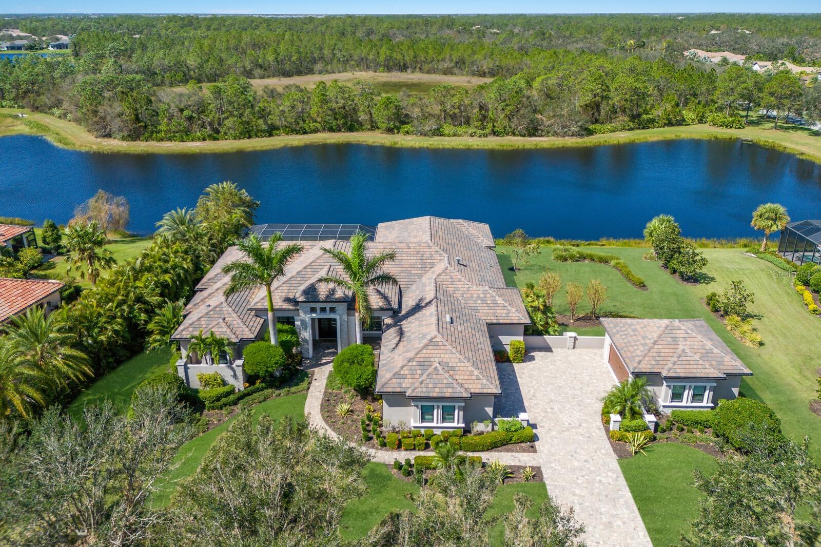 Stellar lake and preserve views on a scenic .70 acre setting in the exclusive and sought-after gated community of The Concession, this four bedroom residence showcases exceptionally planned indoor and outdoor spaces, generously styled detailing throughout, a whole-home GENERATOR, and even a 5 CAR GARAGE. Built 2017, the custom Johnson Homes’ Oakmont plan boasts 4,020 square feet of one-story living and a vastly expansive 1,500+ square feet outdoor living area with abundant open and under cover spaces, pavered deck, full summer kitchen with 2023 grill, PebbleTec heated infinity pool and spa, gas fire table, and a clearview lanai screen.
Seamlessly connecting this resort-styled lanai to the oversized great room, kitchen, and dining areas are the disappearing 10’ IMPACT GLASS doors into an invitingly open space adorned with large scale 24”x48” porcelain tile, soaring ceilings with designer details, intimate wet bar, built-in wine storage and wine refrigerators, and anchored by the designer feature wall with gas fireplace. Chefs and guests alike will delight in the beautifully appointed kitchen with stainless steel Bosch appliances, sprawling Silestone counters, oversized island, wood cabinetry, dovetail soft-close drawers, herringbone patterned backsplash, and a hidden walk-in pantry.
Relax and unwind in the grand-sized owner’s retreat positioned on a private wing, offering two walk-in customized closets, double tray ceiling with crown molding, direct lanai access, and a luxuriously appointed spa-like bath with separate vanities, oversized shower and separate soaking tub. Overnight visitors will be right at home in three additional guest suites, each with walk-in closets, two are ensuite with private full baths, while the third is just off the home’s fourth full bath. Behind double French doors is a voluminous bonus room with stunning coffered ceiling, stone accent wall, and sliders that open to the lanai. Additionally the residence offers a spacious study/den off the owner’s retreat via double entry doors adorned with wide plank wood flooring.
Other highlights of this 6,702 total square foot residence includes impact windows/doors/sliders, whole home generator, central vacuum, two tankless water heaters, solid core interior doors, Crestron smart home system, perfectly sized laundry room with cabinetry and sink, custom built-in bench and cubbies at garage entry, landscape lighting, Eagle textured slate upgraded roof tile, 10’ Mahogany double front doors with 3-point locking system, wood tongue and groove ceilings on front porch and lanai, an oversized attached three-car garage and detached two-car garage with air conditioning unit (both with heavy flake epoxy flooring and insulated impact overhead doors). This captivating residence proudly resides within an exclusive community setting of striking oak hammocks, pines and large lakes, renowned as the home to the World Golf Championship PGA golf tournament in February 2021 and to the World Champions Cup in December 2023, all near dining, shopping, polo, events in the vibrant Lakewood Ranch, Waterside Place, and UTC area, and nearby enough to the enriching culture and beaches of Sarasota.