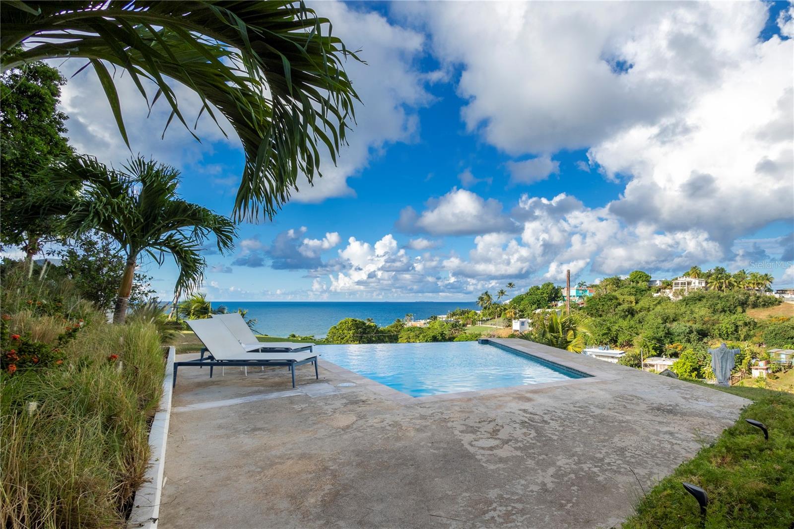 Located in the desirable Bravos de Boston neighborhood, this stunning home offers sweeping mountain and sea views and unforgettable sunsets, all just minutes from the heart of Isabel II and all it’s amenities. Set atop a hill with a north-facing orientation and an “eagle’s nest” vantage point, the property provides privacy, panoramic vistas and refreshing coastal breezes, creating a great home for outdoor island living.
The outdoor terrace, a highlight of the property, features a stunning infinity pool with a vibrant blue-tiled surface that create a glass-like reflection of the changing skies throughout the day. The versatile area includes spaces for lounging, dining, working, as well as a luxurious outdoor shower lined with blue iridescent tiles. The terrace captures the essence of the Caribbean with travertine tiled floors and a unique two-tiered, West Indies style roof that allows light and air to flow freely while still offering ample shade.
Inside, the home is finished with elegant solid stone tile flooring and glass louvers that create a seamless connection between indoor and outdoor spaces. The newly remodeled kitchen features modern finishes, polished cement countertops, and custom PVC cabinetry, for a minimalist design that enhances both style and durability. The open, sun-filled great room stays cool thanks to the natural breezes and has a dedicated living and dining area as well as a work/study. The master bedroom is nestled in the back for privacy and includes a large en-suite bathroom with an outdoor shower. The set-apart guest bedroom is adjacent to a full guest bath and enjoys ocean views. The bedrooms are air-conditioned for added comfort during the warmer summer months.
The landscape is beautifully designed, featuring a mix of palms, including coconut and areca, along with vibrant bougainvillea, frangipani, beach grass, and a meticulously manicured lawn. The property is fully gated for privacy, with a paved driveway for two cars secured by a motorized gate. Additional guest parking is available at the rear, along with an outdoor shower to rinse off after a day at the beach.
Additional features include a propane generator that powers the entire home, a separate laundry room and service closet, a small garden shed, and a pool pump/storage room tucked away for quietness.
This turnkey home is an exceptional find, offering the perfect balance of comfort, privacy, and coastal charm.