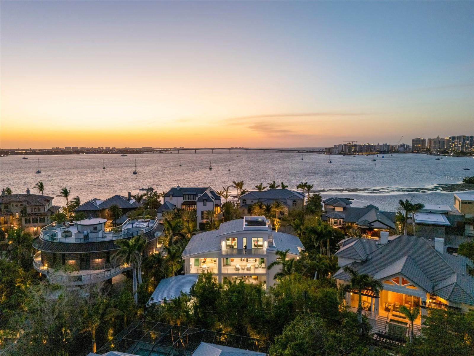 Enjoy the prestige of Harbor Acres, Sarasota’s premier neighborhood, just minutes from downtown and its vibrant Bayfront. This like-new, contemporary estate is more than a home—it’s a statement of intent. Designed for those who value clean lines, modern architecture, and exceptional design, this three-story residence delivers an uncompromising standard of luxury. Step through a dramatic glass entryway into a bright, expansive foyer, where a modern floating staircase and a Vertech 3-stop elevator introduce the home’s refined vertical layout. Immediate access to dual climate-controlled garages with parking for up to 6 cars, finished with epoxy flooring and commercial-grade doors, underscores the home's functionality and thoughtful design. The main living level is centered around a sprawling open-concept great room, ideal for both grand entertaining and intimate living. Gallery-style walls, 12-foot multi-stack sliders, and floor-to-ceiling glass invite in abundant natural light. The chef’s kitchen is a true showpiece, outfitted with Miele appliances, sleek Dekton countertops, Westwood custom cabinetry, and a full butler’s pantry with Sub-Zero refrigeration. An informal dining area and TV lounge extend naturally outdoors to the covered summer kitchen—complete with outdoor grill and thermally modified tongue-and-groove ceilings—creating a seamless indoor-outdoor living experience. The north wing of the main floor is dedicated to a dual-owner’s suite: two bedrooms—one currently used as a home office—joined by a spa-like bathroom featuring floating vanities, dual rain showers, a soaking tub, and specialized Italian PRESOTTO closets. A discreet laundry room and appliance garage further enhance convenience and daily efficiency. Upstairs, a flexible landing space connects a private guest suite with a library and media room—perfect for remote work, relaxation, or extended stays. An additional full bath serves this level, maintaining privacy and practicality. Beyond the main residence, the backyard becomes a private resort. A sleek pool and integrated spa are flanked by lush bamboo privacy landscaping and lead to a standalone guest house featuring a full kitchen and bath—ideal for guests, in-laws, or live-in support. A striking exterior staircase links all levels while maintaining architectural harmony. State-of-the-art features include a top-of-the-line Crestron smart home system, whole-house generator, tankless water heaters, an at-home gym, and secure gated entry. Crowning the estate is a private rooftop terrace offering panoramic 360° views of Sarasota Bay, the Ringling Bridge, Longboat Key, Siesta Key, and downtown Sarasota—an unmatched setting for sunset entertaining or tranquil reflection. This is more than a home. It’s a lifestyle of prestige, privacy, and purposeful design—for those who live without compromise.