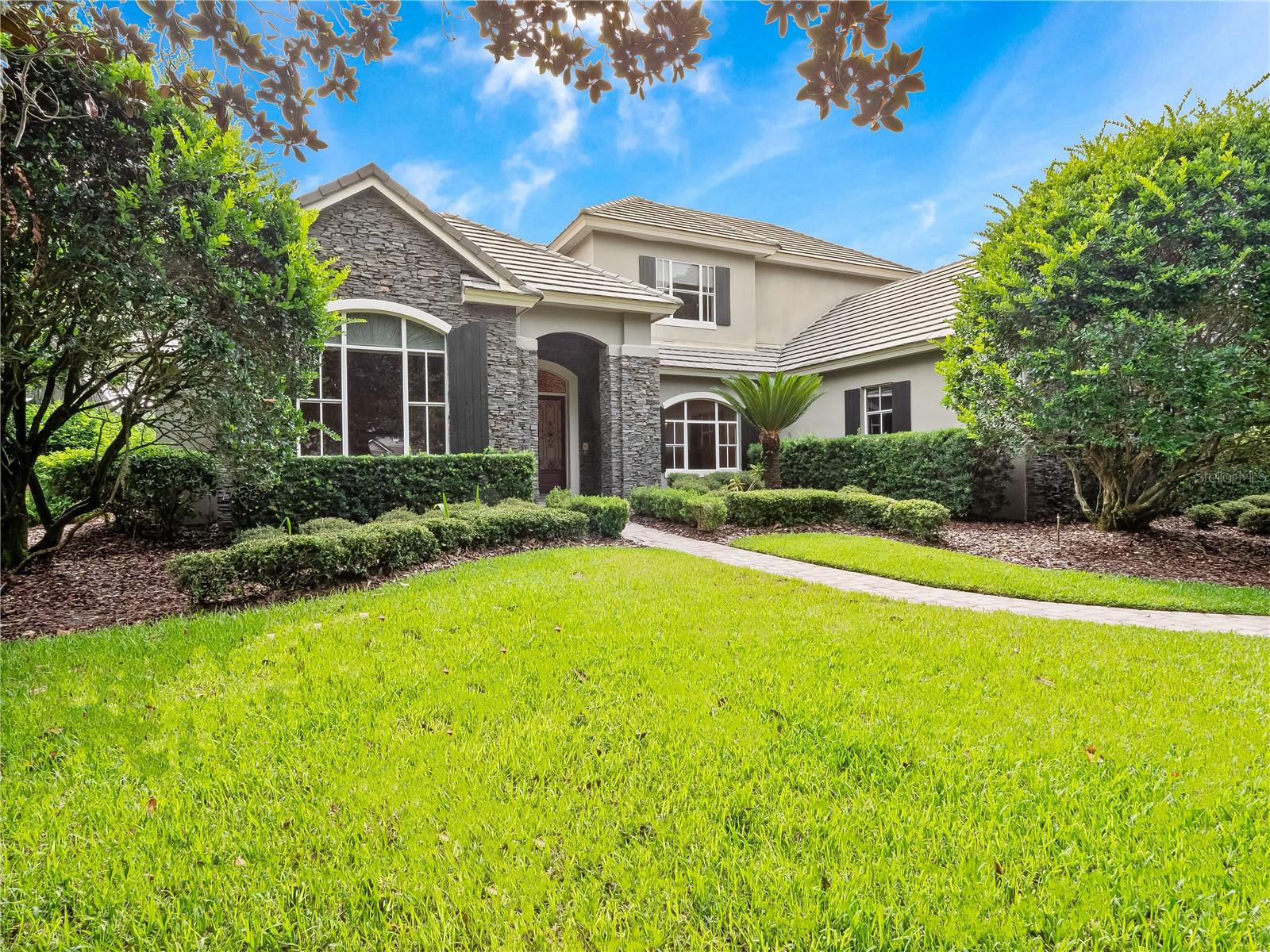 One or more photo(s) has been virtually staged. Welcome to this beautiful residence in the highly sought-after Alaqua Lakes community where thoughtful design and timeless elegance meet everyday comfort. Nestled on a private ½-acre lot with a spacious backyard, this home offers exceptional curb appeal with its inviting exterior elevation and striking stone accents. Inside, you’ll find soaring ceilings, quality finishes and generous living spaces that blend sophistication with warmth and character.
At the heart of the home lies the gourmet kitchen designed to inspire both everyday living and elevated entertaining. Overlooking the family room and the bright and airy sunroom, the kitchen is a true gathering space filled with natural light and seamless functionality. Just steps away are the formal and casual dining areas, a 1,200-bottle climate-controlled wine room and a butler’s pantry making hosting effortless and stylish.
A dedicated office with custom built-ins and a cozy fireplace offers the ideal setting for remote work or a quiet study. The grand primary suite, located on the first floor, serves as a private retreat with a spa-like en suite bath featuring split vanities, granite countertops, dual sinks, a whirlpool soaking tub, and two spacious walk-in closets. Upstairs there are three generously sized bedrooms and two full bathrooms that provide comfortable accommodations for family or guests along with plentiful storage throughout.
Set within a premier golf course community, residents enjoy world-class amenities including clay tennis courts, a resort-style pool, a 24-hour guard-gated entrance and enhanced community security. Ideally located near Central Florida’s best attractions, upscale shopping and dining, major highways, and international airports, this exceptional home delivers a rare combination of luxury, privacy, and lifestyle.