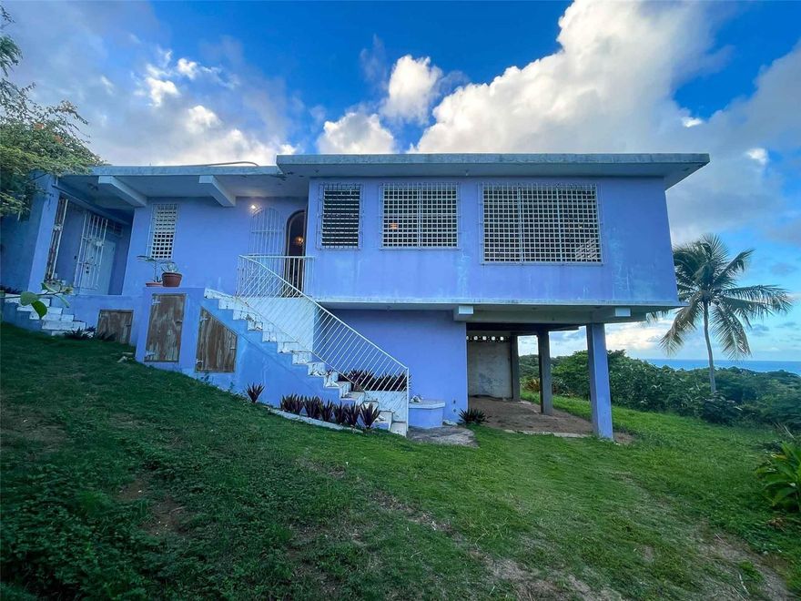 Casa Verde is a 1-bedroom, 2-bath concrete home nestled into the green hills above Monte Santo on Vieques Island, Puerto Rico. Perched at the end of a quiet road and surrounded by mature gardens, palm trees, and pastureland, this property offers both privacy and panoramic views stretching from the north coast of Vieques to Culebra, mainland Puerto Rico, and the Caribbean Sea.

Set on a fully fenced half-acre parcel, Casa Verde features a two-level layout with an elevated main floor and ground-level covered parking and storage. The home includes a unique opportunity to expand upward, with an exterior staircase leading to an unfinished rooftop terrace—offering the potential to create a spectacular third-floor deck or additional living space.

The main living area is light-filled and open, with tall ceilings, abundant windows, and an airy layout that connects the living room, dining area, and kitchen. French doors open to a covered balcony that captures the trade winds and delivers daily views of ferry boats, sunrises, and sunsets.

The kitchen features a gas stove, decorative tile work, open shelving, and a central island ideal for food prep, crafts, or casual dining. A wall of windows floods the space with natural light. Painted concrete floors throughout add durability and character.

The primary bedroom accommodates a king-sized bed and includes an ensuite bathroom with a glass block shower and direct access to a private back patio with an outdoor shower and bathtub—perfect for rinsing off after the beach or enjoying a soak under the stars. A laundry closet is conveniently located within the primary suite.

A "second" bedroom, separated from the main living area by a partial wall, includes a large closet and potential for privacy improvements. The home also includes a bonus room/office space that could serve as a third sleeping area, and a second full bathroom accessible to guests and the secondary bedroom.

Surrounding the house is a lush and carefully curated garden, filled with tropical plants and mature palms that frame the panoramic views. Located at the top of the hillside neighborhood above Monte Santo, the home enjoys a serene setting with minimal street traffic and easy access to both Isabel Segunda and the island’s north shore beaches.

Whether you’re looking for a peaceful full-time residence, a Caribbean vacation home, or a flexible island retreat with room to grow, Casa Verde offers comfort, style, and island charm in equal measure.