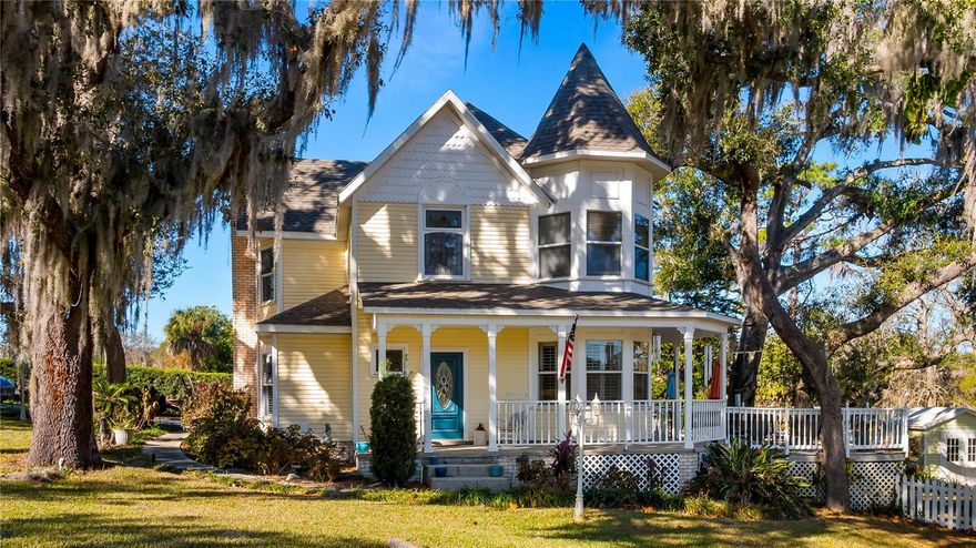 Welcome to this beautifully designed Victorian-style home built in 1992, offering over 2,088 square feet of living space and nestled on just under half an acre beneath majestic oak trees in desirable Clermont, Florida. The front porch warmly welcomes your guests and wraps around to the back porch and deck, creating the perfect setting for relaxing and entertaining. Ideally located near downtown Clermont between Lake Minnehaha and Lake Minneola—part of the Clermont Chain of Lakes—this home is walking distance to a local park and enjoy a scenic view of Sunnyside Lake.
A welcoming foyer greets your guests and sets the tone for the charm found throughout the home. This residence features three bedrooms and two and a half baths with a thoughtfully designed layout. The large kitchen overlooks the family and living area and includes all appliances, two pantries, and convenient access to the garage, while offering beautiful views of the back porch, deck, and spacious backyard—perfect for everyday living and entertaining. The bright and open living room showcases plantation shutters and a cozy gas fireplace, and the formal dining room, located just off the kitchen and foyer, also features plantation shutters and provides an inviting space for gatherings and special occasions.
Upstairs, the spacious primary bedroom is highlighted by the home’s signature “witch’s hat” turret, creating a charming space ideal for reading and relaxing while enjoying views of Sunnyside Lake. Master offers two walk-in closets.  Two additional guest bedrooms and a full bath complete the second level, offering comfortable accommodation for family or visitors. Garage has a 10’ x 10’ office that is under air.  Recent updates include a new roof in 2023 and a new A/C in 2020.  Hot Water Heater 2017. Sidewalk recently added by the city.  Storage Shed in backyard.  Picket fenced backyard.  With its lush landscaping, classic Victorian character, wraparound porch, and prime location near lakes, parks, and downtown Clermont, this unique home offers a rare opportunity to enjoy timeless charm paired with modern comfort.