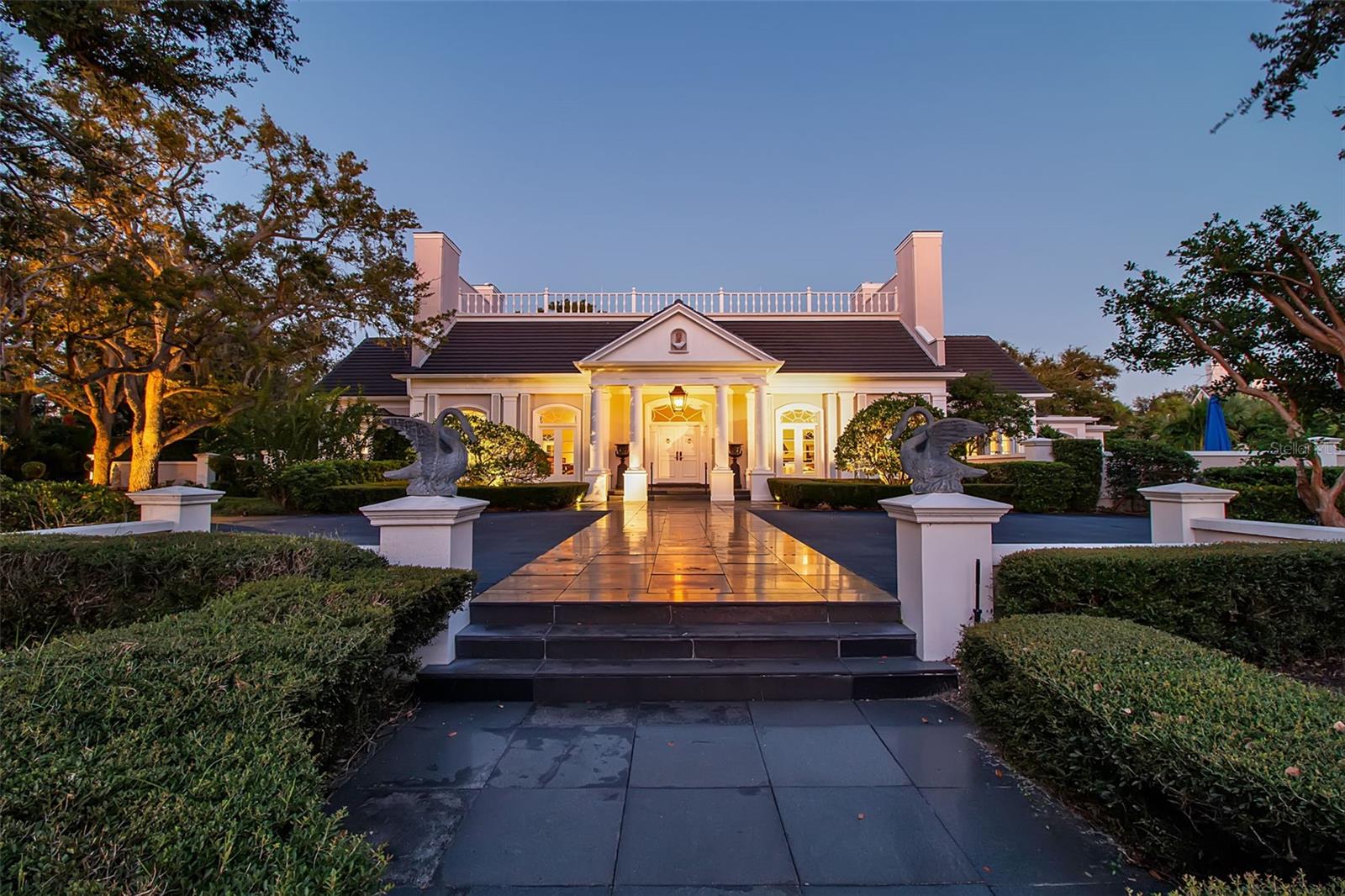 A grand estate where timeless design meets lakefront tranquility, 101 Osprey Point Drive offers refined living within the private gates of The Oaks Bayside. Designed by architect Bo MacEwen, this one-of-a-kind home spans more than 5,000 square feet, all cleverly positioned on nearly an acre with 150 feet of lakefront. Situated on two of the community’s most desirable lots, the residence balances architectural distinction with thoughtful updates that ensure comfort and livability. An oversized circular drive paved in granite tile leads to a stately double-door entrance. Inside, the foyer sets the tone with soaring ceilings, hand-crafted moldings, chandeliers, and a white onyx floor with marble inserts. Spaces include an oversized living room with dual fireplaces, a crystal chandelier, and multiple sets of French doors opening to the lakefront patio. The dining room, anchored by another fireplace and framed by custom casings and built-in shelving, is ideal for grand entertaining. A domed ceiling sitting room with French doors to the patio adds an intimate space for cocktails or conversation, while the custom bar and wine room, with etched glass cabinetry, stone counters, and display shelving, enhances the home’s appeal for gatherings. The kitchen, completely remodeled in 2019, is a showcase of craftsmanship with UltraKraft® cabinetry, custom soffit lighting, Corian countertops, and a center island with breakfast bar. Luxury appliances include a Sub-Zero refrigerator, Wolf gas cooktop, wall oven, microwave, warming drawer, two KitchenAid dishwashers, and a Frigidaire wine fridge. A butler’s pantry provides additional storage, while the sunny breakfast nook overlooks lush gardens. Family and gathering rooms carry the same sense of scale, with dramatic fireplaces, intricate moldings, and detailed ceiling treatments that flow seamlessly for everyday living or large-scale entertaining. The primary suite is a true retreat with a spacious sitting area, custom closets, and a well-appointed bathroom that was newly remodeled in 2025 with spa-inspired finishes. Three guest suites, each with updated full baths (2021 and 2025), provide privacy and comfort. One is a suite with a bathroom near the laundry room, which offers flexibility for live-in staff or extra accommodation. Outdoors, the home embraces its setting with an open pool, expansive deck, and cabana surrounded by manicured hedges and mature landscaping. The views are equally compelling, with mature gardens and community reflecting pond in the front and sweeping lake vistas in the back. Additional highlights include a three-car garage, refinished hardwood floors (2020), and a new Daikin 5-ton AC system installed in 2022. Ownership at The Oaks Club brings an unmatched lifestyle, with two championship golf courses, tennis and pickleball courts, croquet, a fitness center and spa, three on-site restaurants, and a community dock on Little Sarasota Bay. Elegant yet inviting, 101 Osprey Point Drive stands as a rare opportunity to enjoy lakefront living with resort-style amenities in one of Sarasota’s most established private enclaves.