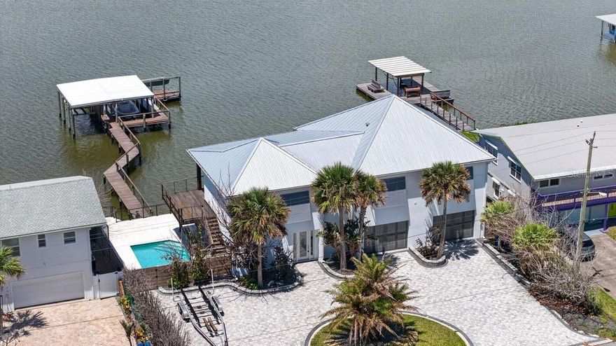 Nestled on 100 feet of pristine Intracoastal Waterway frontage in the coveted Bethune Beach neighborhood of New Smyrna Beach, this extraordinary custom-built coastal estate delivers an unrivaled blend of luxury, craftsmanship, and waterfront lifestyle just steps from a private beach walkover and the untouched beauty of Canaveral National Seashore. Completed less than 3 years ago with no expense spared, this fully furnished turnkey masterpiece showcases breathtaking panoramic river views with no homes across the water, offering unmatched privacy and spectacular sunsets. Elevated high above the water with a seawall, multi-tier retaining walls, and lush tropical landscaping, the property features an IPE dock complete with water, cleaning station, floating dock, and 2 covered boat lifts. A circular paver driveway accommodates up to 10 vehicles in addition to the oversized 36' wide two-car air-conditioned garage featuring tiled floors, 10' ceilings, custom garage doors with side-mount openers, wet bar with Hoshizaki commercial ice machine, Thermador wine cooler, 2nd laundry, and massive storage room for all your gear. Inside, dramatic architecture and refined modern design immediately captivate with smooth walls, a stunning underlit floating IPE staircase with cable rails, and a commercial-grade elevator with auto-glass gate offering unforgettable water views as it ascends to the main living level. The first floor hosts 2 luxurious riverfront guest suites and direct access to the resort-style pool with travertine decking and covered lanai. Upstairs, soaring vaulted ceilings frame the expansive riverfront living, dining, and entertaining spaces, all opening through walls of impact sliders to covered and open-air IPE-clad terraces with electric sunshades overlooking the water. The showstopping chef’s kitchen is truly one of a kind, appointed with Sub-Zero refrigeration and freezer drawers, Viking gas range with custom hood, double Thermador wall ovens, Bosch microwave drawer, prep sink, quartz countertops, and wine cooler. The lavish primary suite offers double French door entry, endless water views, custom walk-in closet, and an extraordinary spa-inspired bath featuring Brizo rain heads and body jets controlled by Moen digital panels, quartz vanities, illuminated mirrors, soaking tub, and smart Brondell toilet. A massive second-floor laundry room with dual access from both the primary suite and kitchen adds exceptional everyday conveniencen. Quarter-sawn oak flooring spans the second level while oversized designer tile flows throughout the lower level and baths. Additional luxury features include aluminum metal roofing, hurricane impact windows and doors, concrete beam-and-column construction with reinforced concrete floors and masonry exterior, Aquasana whole-home filtration, hybrid water heater, Honeywell security system with cameras, recessed LED lighting, electric blinds for west-facing windows, Moen digital shower systems, smart toilets throughout, built-in wifi expanders, surround sound, and premium Arhaus furnishings included, in addition to Polywood exterior seating. Located just minutes from groceries, fine and casual dining, shopping, and vibrant nightly entertainment, this is a rare opportunity to own one of Bethune Beach’s most exceptional modern waterfront estates. ALL INFORMATION IS ASSUMED ACCURATE BUT NOT GUARANTEED. ALL MEASUREMENTS ARE APPROXIMATE. BUYERS AND THEIR AGENTS SHALL VERIFY INFORMATION DEEMED IMPORTANT TO THEM.