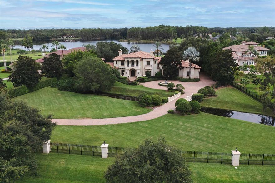 Welcome to this private lakefront estate in the prestigious gated community of Steeplechase in Odessa. Positioned on more than three acres, this custom residence offers 6,749 square feet with five en-suite bedrooms and six full baths. A tree-lined paver drive leads to the stately façade, where arched windows, manicured hedges and a circular driveway with grand fountain welcome you in. Inside, soaring ceilings and marble floors set a tone of classic refinement. Designed for entertaining, the home features formal living and dining areas, a chef’s kitchen with Wolf range and Sub-Zero refrigerator, and an open layout that flows seamlessly from indoors to out. Retreat to the expansive first-floor primary suite, complete with a fireplace, flex room, and spa bath. Guests will enjoy their space in bedrooms with en suites, with one downstairs and three more on the upper level. A stately office with rich wood paneling, a wine cellar with climate control, and a masculine lounge/man cave elevate the luxury lifestyle even further. Outside, enjoy four distinct outdoor living areas: a columned lanai, a sparkling pool and spa, and a fully equipped summer kitchen — all surrounded by lush, mature landscaping and privacy. Additional luxuries include a central vacuum system, dog washing station, four-car garage with two EV chargers, and a whole-house generator.