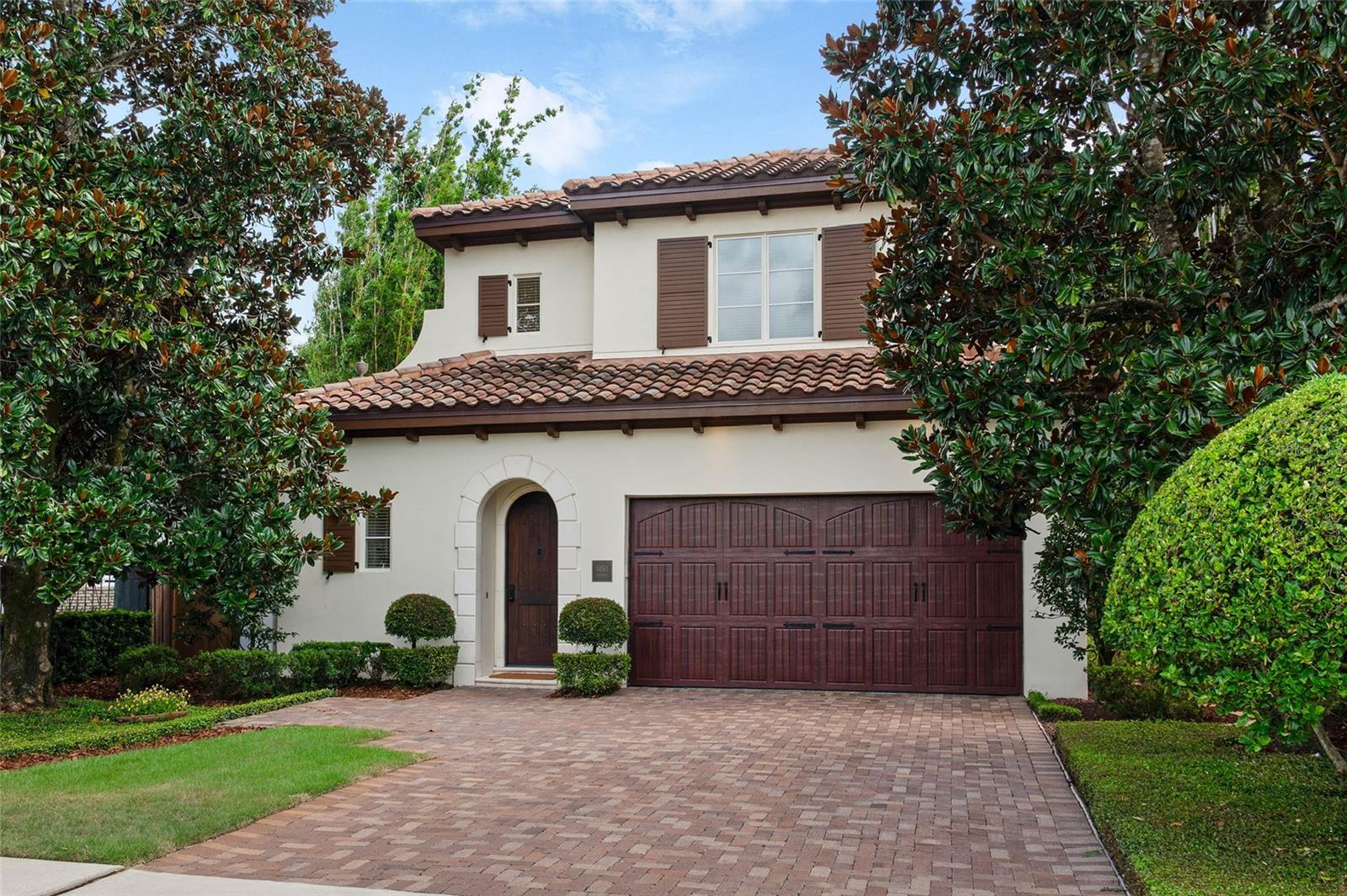 Located in the coveted Tree Streets just a block and a half from Lake Knowles, this timeless Mediterranean home offers three bedrooms, three and a half bathrooms, and just under 2,000 square feet of beautifully finished living space framed by mature magnolias and manicured hedges. A brick driveway leads to a two-car garage with carriage-style doors, while a covered entry showcases a solid arched wood front door with classic peep hole, setting the tone for the craftsmanship inside. Step through the entry and discover a hidden laundry room with dark wood cabinetry, marble countertops, subway tile backsplash, and front-loading washer and dryer, along with a sophisticated powder bath featuring a floating marble-topped sink, wall-mounted chrome faucet, integrated mirror lighting, and detailed molding. Just beyond is a flexible room that functions as a dining room or formal living space, and through the next doorway the home opens to a large kitchen and great room designed for everyday living and entertaining. The kitchen combines dark wood perimeter cabinetry with accent glass fronts, a creamy white island with storage on both sides, marble countertops, and stainless appliances including a GE Monogram gas cooktop with vent hood, convection oven, refrigerator, dishwasher, and LG under-counter microwave, all anchored by a deep single-barrel undermount sink with extendable faucet. The great room features a gas fireplace with mosaic tile surround, clerestory windows, extensive crown molding, and hardwood floors, with new Anderson French doors that open to the private backyard where brick decking frames a cocktail pool with water features by Signature Landscape, surrounded by mature greenery and magnolias. The primary suite, tucked on the back right wing, also enjoys French doors to the pool terrace and includes new carpet, a customized floor-to-ceiling walk-in closet, and a bath with travertine floors, sunken glass-enclosed shower with built-in shelving, creamy white vanity topped with marble, satin nickel fixtures, dual Kohler undermount sinks, and mirrored walls with crown molding. Upstairs, two spacious bedrooms each feature ensuite baths and customized closets—the front with a built-in desk, marble chicklet tile flooring, creamy cabinetry, and tub/shower combo, the second with a walk-in closet and a bath finished with travertine flooring, a slightly larger vanity with consistent cream cabinetry, marble top, and glass-enclosed shower. Additional highlights include hidden storage under the stairs, unique iron stair railing, 8’ solid wood interior doors, 10’ ceilings, and block construction, while recent updates such as an Italian tile roof (2020), newer garage door and opener, and new HVAC air handler provide peace of mind. Just a short walk to the YMCA, Phelps Park, and Interlachen Country Club, 1450 Chestnut Avenue combines Mediterranean charm, thoughtful updates, and a sought-after Winter Park location for a lock and leave lifestyle with ease.