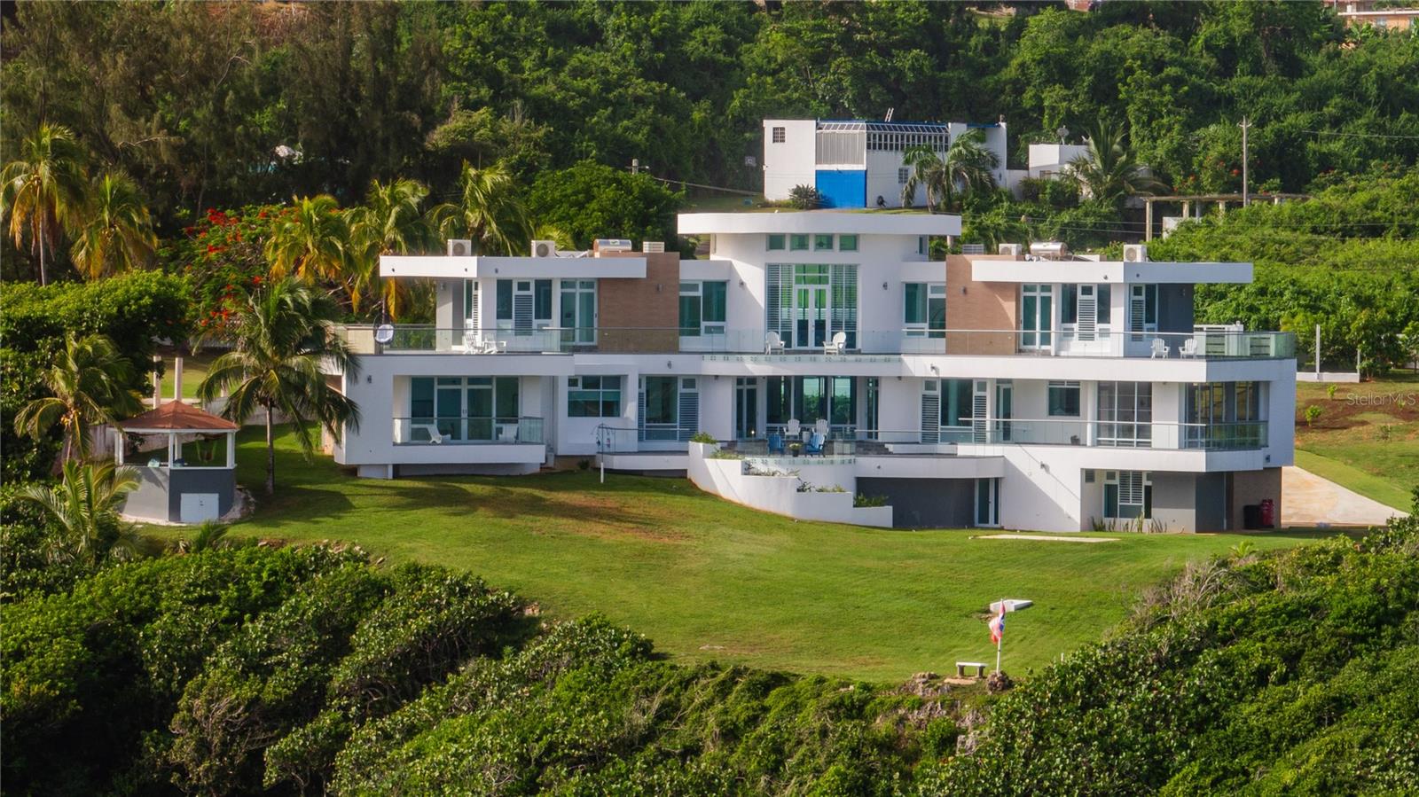 Oceanfront Estate with Panoramic Views in Quebradillas’ Terranova Neighborhood
Welcome to a truly extraordinary coastal estate in the sought-after Terranova neighborhood of Quebradillas, Puerto Rico. Positioned 170 feet above sea level, this architectural masterpiece offers breathtaking, unobstructed ocean views from every single room—including all bedrooms, living areas, kitchen, formal dining, home office, and even the guest house.
Completed in 2025, the main residence spans 6,515 sq. ft. across three beautifully designed levels. The entry level (second floor) features a stunning open-concept kitchen with granite countertops, a center island, custom cabinetry, a breakfast room, and a walk-in pantry. The living room, home office, and kitchen flow seamlessly into a wraparound terrace, while the screened formal dining room offers panoramic water views for a one-of-a-kind entertaining experience.
The primary suite is a private retreat with a custom walk-in closet and an expansive spa-style bathroom complete with double vanities and a large walk-in shower with dual shower heads. Upstairs, two additional bedrooms each feature private en-suites, walk-in closets, and access to shared ocean-view terraces. The third floor also houses a dramatic game room with soaring 20-foot ceilings, a wet bar, and uninterrupted views of the Atlantic.
The ground level includes a three-car garage, a large storage room, a secondary laundry area, and a full bathroom with direct backyard access.
The guest house, built in 2023, offers 1,200 sq. ft. of thoughtfully designed space with two bedrooms and two bathrooms, including an ensuite in the primary bedroom. The living, dining, and kitchen areas all feature stunning ocean views, and the home is fully equipped with independent electric and water meters, as well as its own septic system.
This estate includes ceiling unit A/Cs in main living areas, mini splits in each room and closet, a concrete privacy fence with electric gate, and infrastructure ready for solar panel installation. Additional amenities include an 18kW GENERAC® propane generator with automatic transfer switch and 200 amp service, a 600-gallon water cistern with a 220V pump and pressure tank, and solar water heaters across both homes. And for nature lovers, the estate offers front-row seats to seasonal whale watching from both homes—brand-new binoculars included with purchase.
Located just 30 kilometers from Aguadilla International Airport and free of HOA or community restrictions, this oceanfront estate is a rare opportunity to own a private slice of paradise where luxury, comfort, and panoramic views converge.