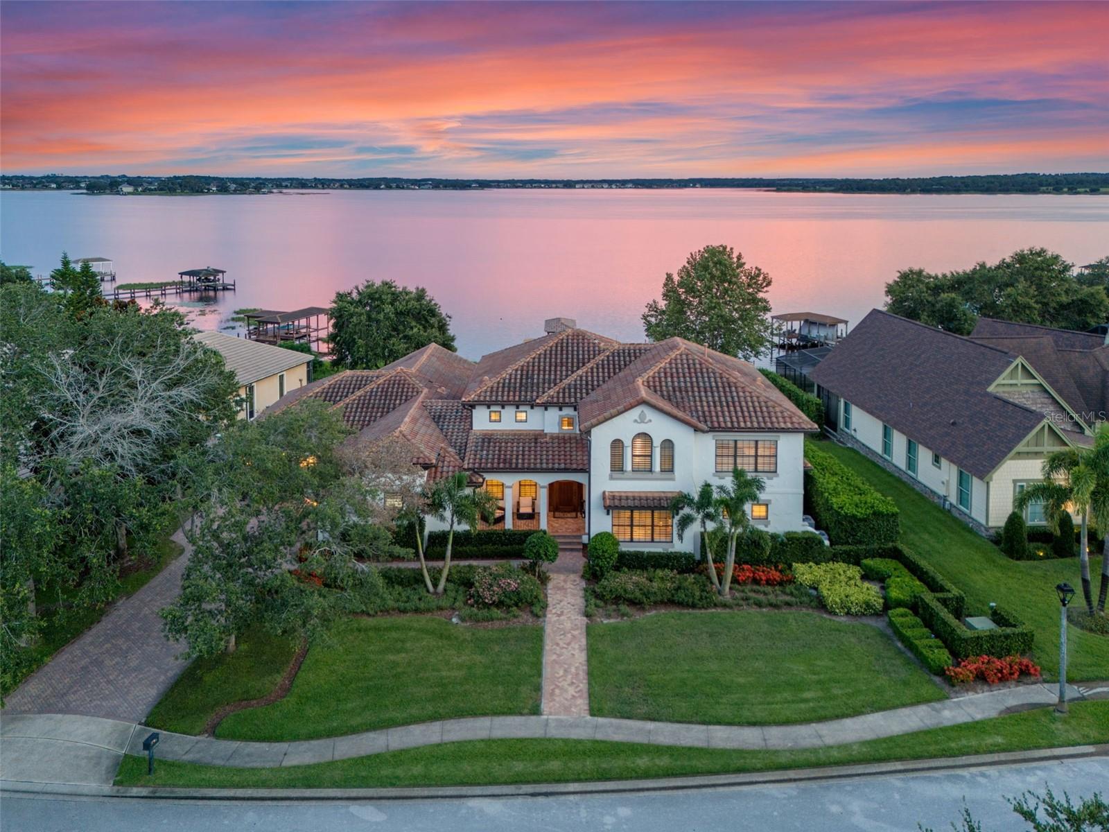 Extraordinary lakefront estate, where culinary excellence meets waterfront paradise. Immerse yourself in the ultimate lakefront lifestyle at this breathtaking direct waterfront estate on Johns Lake, where every morning begins with sunrise reflections dancing across 2,500 acres of pristine water and every evening concludes with golden sunsets painting the sky from your private south-facing sanctuary. This magnificent 4,800=-square-foot home in a gated community represents the pinnacle of Florida waterfront living, blending indoor luxury with endless aquatic adventures right outside your door. This amazing gourmet kitchen showcases stunning one-piece stone countertop that flows seamlessly across the expansive workspace in a single level, creating visual drama and the kitchen's design brilliance extends to its abundance of new custom cabinetry, meticulously crafted to maximize storage and style, with a cleverly hidden walk-in pantry. A charming breakfast nook features built-in bench seating positioned to capture the lake view. The built-in bar area elevates entertaining with its ice maker, wine refrigerator and elegant bottle storage/display options, allowing hosts to craft cocktails. A formal dining room is spacious enough to comfortably accommodate 10 guests beneath a gorgeous statement light fixture. Your waterfront lifestyle extends far beyond the home's solid concrete walls to encompass the entirety of Johns Lake where there are weekly fishing tournaments, peaceful kayaking or paddleboarding, water skiing and wakeboarding and leisurely pontoon cruises. Your new private dock, complete with boat lift, provides immediate access. This five-bedroom, three full and two half-bath residence offers dual primary suite luxury with one retreat on each level. The main floor primary suite captures gorgeous lake views through expansive windows, features a romantic corner fireplace elegantly set in travertine and has a generously sized walk-in closet with custom organizers. The bath presents a freestanding soaking tub, a beautiful glass-enclosed shower, dual vanities and a built-in makeup desk. The fifth bedroom on the main level provides flexibility as either guest accommodation or home office. The elegant spiral staircase, adorned with wrought iron and wood railings is dramatically illuminated by an amazing multi-level drop pendant light fixture. Here, an incredible theater room with comfortable seating transforms movie nights while the huge upstairs family room offers space for a second family room. The second primary suite offers those same lake views, has a large walk-in closet and gorgeous bath. Additional guest bedrooms, including a rear bedroom that captures amazing vistas, ensure comfortable accommodation for family and visitors. Large laundry room with custom cabinetry and utility sink, garage entry area includes built-in seating and storage solutions for shoes, backpacks and everyday essentials. The three-car garage, crown molding and new flooring throughout the home create a cohesive atmosphere of refined elegance. Fantastic location as the Florida Turnpike is three minutes away, dramatically reducing commute times. This remarkable lakefront estate represents more than a home. It embodies a lifestyle where gourmet culinary experiences unfold against a backdrop of endless water views, where morning coffee transitions seamlessly into afternoon water sports and where every sunset provides daily reminder of life's extraordinary possibilities.