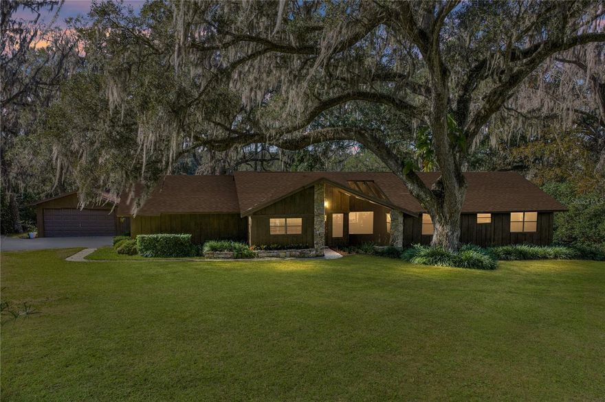 Escape to nearly 3 acres of pure Florida tranquility in this peaceful, rustic-style lakeside retreat offering 180 feet of sparkling water frontage, sweeping views, a 2024 roof, a 2025 AC, and a lush tropical landscape dotted with grand, mature shade trees. Though you’re just minutes from downtown Dade City, this 3 bedroom, 2 bath, 4 car garage property feels worlds away, your own private lakeside getaway. Step inside to a warm, welcoming layout where spacious, dedicated rooms flow easily from one to the next. The striking 24x15 living room is a true showpiece: rich wood paneling, wood-beam accents, a floor-to-ceiling stone fireplace with wood mantle, and quadruple recessed sliders that open wide to the screened porch and serene lake views. A passthrough window connects the living room to the kitchen and dining area, keeping the heart of the home connected. And what a kitchen it is- an dream for anyone who loves to cook. Joined seamlessly with the dining room for a genuine eat-in experience, it offers a gas range, refrigerator, dishwasher, generous prep and storage space, a built-in breakfast bar, and a convenient desk nook. A sliding door leads directly to the open deck overlooking the water, perfect for morning coffee or evening unwinding. Just off the kitchen, the garage and laundry access make daily routines effortless, from groceries to chores. The private bedroom wing features three comfortable bedrooms with plush carpeting and ceiling fans. A full guest bath with a tub/shower combo sits conveniently in the hallway. The primary suite stands out with a spacious walk-in closet and an ensuite bath complete with dual sinks and a walk-in shower. Outside, the possibilities are endless. Enjoy the open deck, screened porch, sprawling yard, or the dock that puts you right on the water. There’s an attached 2 car garage plus a detached 2 car garage, ideal for projects, storage, or extra enclosed parking. Spend your days soaking up sunshine, tinkering in the workshop spaces, heading into town for local events, or simply relaxing while the lake breeze drifts through the trees. This is the kind of place people dream about- private, peaceful, and filled with natural beauty. This is lakeside living at its best, schedule your private showing today!