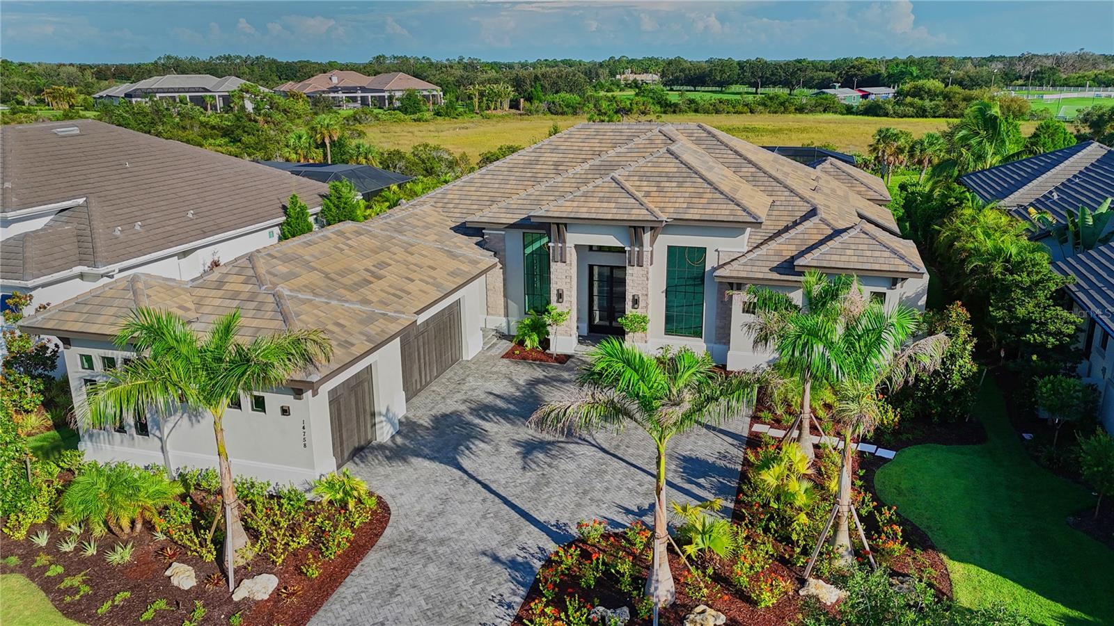 Transitional West Indies Custom Home by John Cannon Homes.
Experience the perfect blend of West Indies architecture with contemporary influences in this brand-new, never occupied custom residence. From the moment you arrive, huge tandem windows, a striking 10-foot custom iron entry door and a symmetrical façade draw your eyes straight through to the breathtaking rear views. Inside, the cathedral ceilings with scissor beams in the great room flow seamlessly to the main lanai and through the expansive pool enclosure. 10-foot accordion stacking doors topped with transom windows open the home to true indoor–outdoor living. A gas fireplace with cultured stone surround and mantle creates a warm focal point, while a climate-controlled wine room with illuminated agate stone panels adds an element of artistry. The home offers both a primary kitchen and a chef’s prep kitchen, each outfitted with high-end appliances. Natural light floods the space through an interior/exterior window and skylights. Two secondary bedrooms each feature en suite baths and walk-in closets. A versatile bonus room includes a disappearing corner door to extend entertaining space to the outdoors. In the owner's wing, you’ll find dual owner’s baths and closets—each closet with LED pin lighting and custom built-in cabinetry. A private owner's courtyard with outdoor shower and composite decking creates a resort-like retreat.
The rear lanai is fully equipped with an outdoor kitchen and island, overlooking the stunning pool area enclosed by a clear-view screen and colonnade. The pool features shellock pavers, a large sun shelf with cascade bubbler, in-pool lounge chairs, and a floating fire pit with mirrored granite edges. The negative-edge spa with glass waterline tile completes the scene. This smart home comes pre-wired for electric window treatments, and includes an integrated camera system, home music, and more—delivering luxury, comfort, and technology in one extraordinary package.