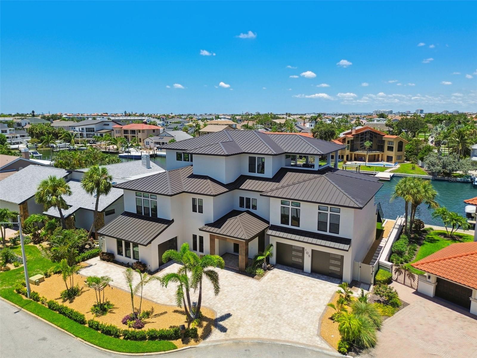 A modern coastal masterpiece, thoughtfully engineered for enduring elegance and the ultimate waterfront lifestyle. Constructed totally custom in 2022, offering 4 bedrooms, 4 full baths, 2 half baths, over 5,500 sq ft of air-conditioned living space, and more than 10,700 sq ft under roof — including expansive patios and a 3,500 sq ft air-conditioned garage with a professional quality gym on the ground level. Designed for long-lasting ownership and true peace of mind, the home is constructed with concrete block on the first level, a ZIP wood system for upper floors, over 59 pilings tied to the full footer system, hurricane-impact PGT windows and sliders, spray foam insulation on all exterior walls and roof, plus batt insulation for interior sound reduction. An aged bronze standing seam metal roof, whole-home Generac generator, twin tankless propane water heaters, and a 500-gallon buried propane tank add durability and resilience. Inside, no detail was overlooked — European oak wide-plank floors run throughout, complemented by Porcelanosa tile in all bathrooms and main laundry. Every door features Baldwin solid brass handles. The gourmet kitchen is outfitted with Sub-Zero and Wolf smart appliances, dual 11-ft islands with waterfall edges, custom Busby cabinetry with accent lighting, a separate pantry, and a dedicated coffee bar. All countertops throughout are Cambria quartz with lifetime chip repair. The primary suite is a true retreat with a private office and an additional flex room along with a spa-like bath featuring a huge no-threshold shower with linear drain, freestanding tub, custom vanities, built-ins, and electric privacy screens. The home includes a SONOS wireless sound system, Lutron Caseta smart dimmers, and automated Hunter Douglas window treatments throughout. Outdoor living is resort-inspired with multiple lanais and decks finished in ¾” porcelain tile, a PebbleTec pool with white travertine coping, dual in-pool spas with Pentair automation, Shawgrass artificial turf, triple built-in umbrellas, and a fully equipped dock with a 20,000 lb boat lift, additional lift for smaller watercraft, full-length aluminum walkways, under-dock blue lights, and dedicated power and water at every slip. The oversized garage (up to 10 cars) features a dedicated A/C unit, epoxy floors, custom flake finish, a pro-grade gym with Rogue, REP, and Titan equipment, and a locking storage closet. This is a rare opportunity to own a true architectural gem where every detail — from the structural engineering to the finishes — was designed to deliver luxury, security, and the ultimate waterfront lifestyle. Offered fully furnished and move-in ready, contact us today to arrange your private appointment to discover the pinnacle of modern coastal living!